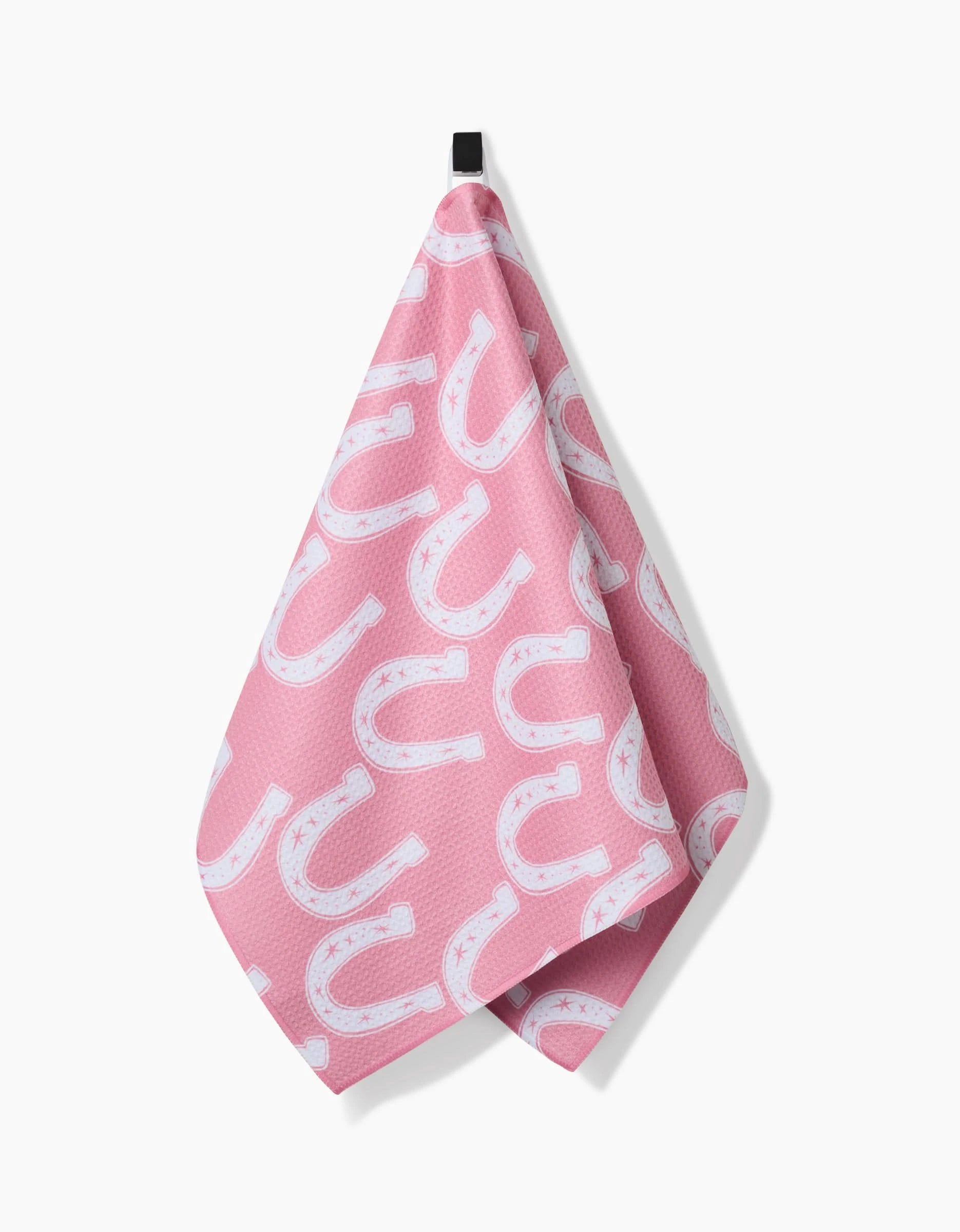 The Lucky Horseshoe tea towel by Aubrey Fairchild is pink, made from recycled materials, features white horseshoes and stars, and hangs from a black loop against a white background.