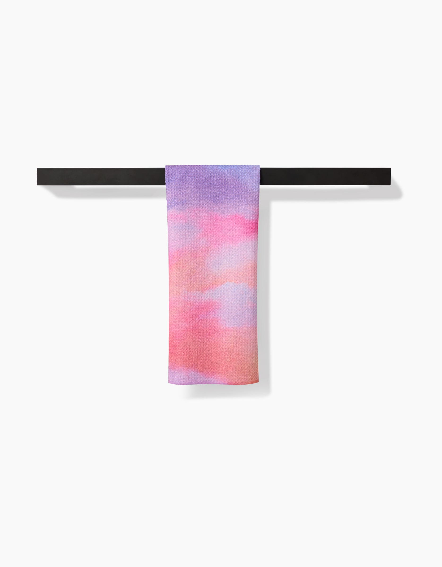The Magic In The Sky tea towel by Stefanie Bales, crafted from soft Geoweave fabric, features a pastel pink, purple, and peach gradient pattern and hangs neatly on a black bar against a white background.