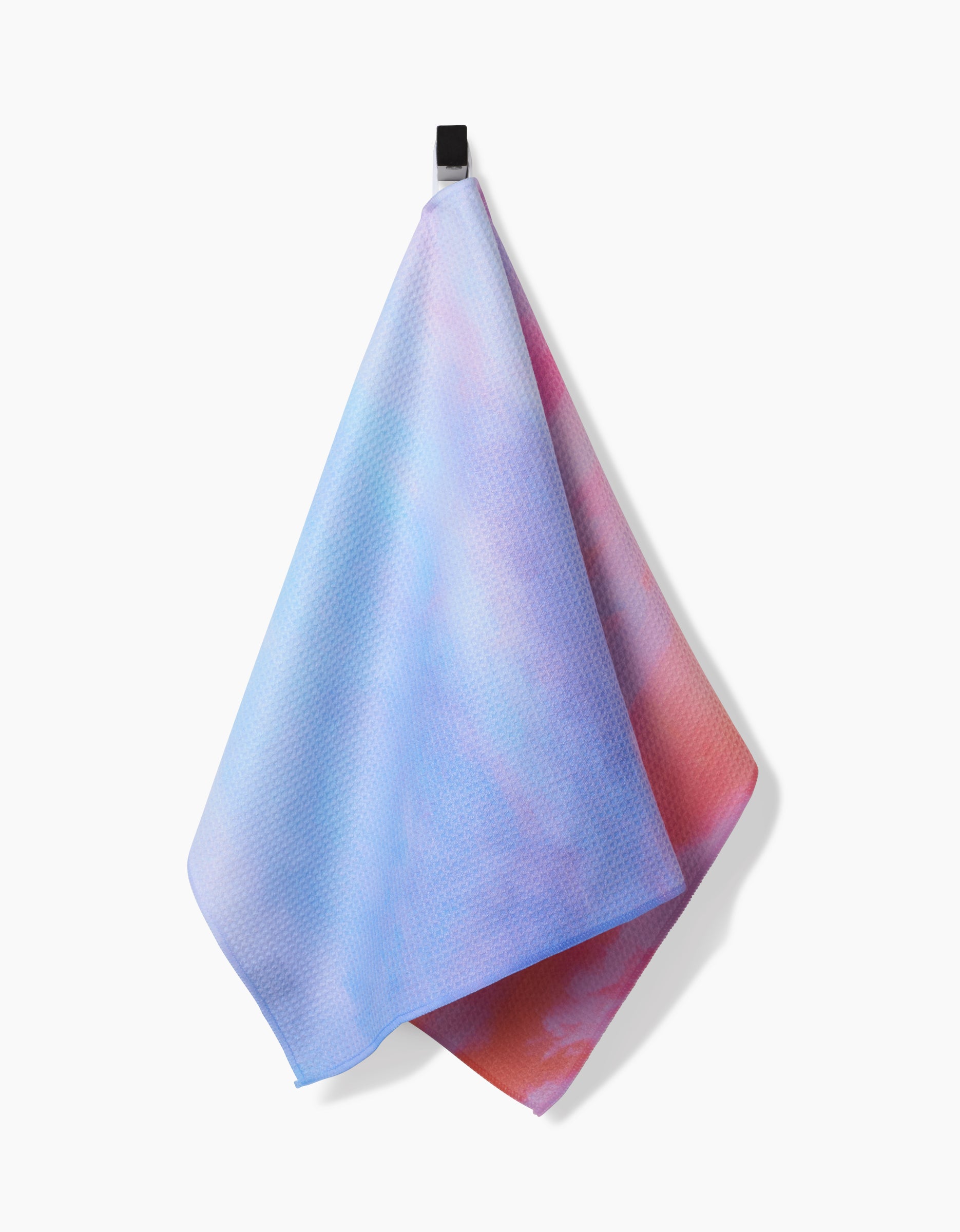 The Magic In The Sky tea towel by Stefanie Bales, featuring pastel blue, pink, and purple Geoweave fabric, hangs from a black hook against a white background.
