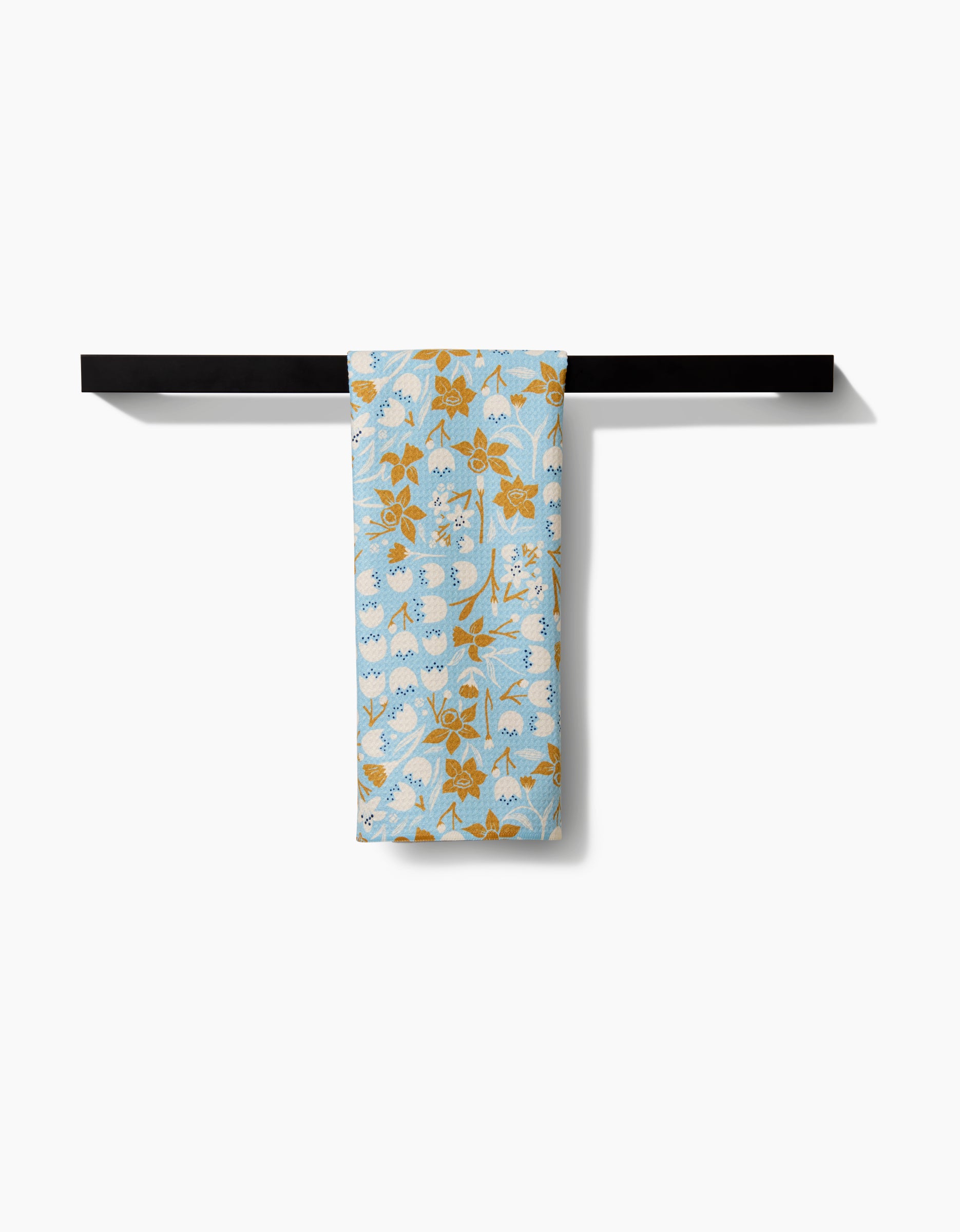 The Elisabeth Wing March Tea Towel, made from recycled materials and decorated with mustard yellow and white flowers, hangs neatly on a black towel rack against a plain white background.