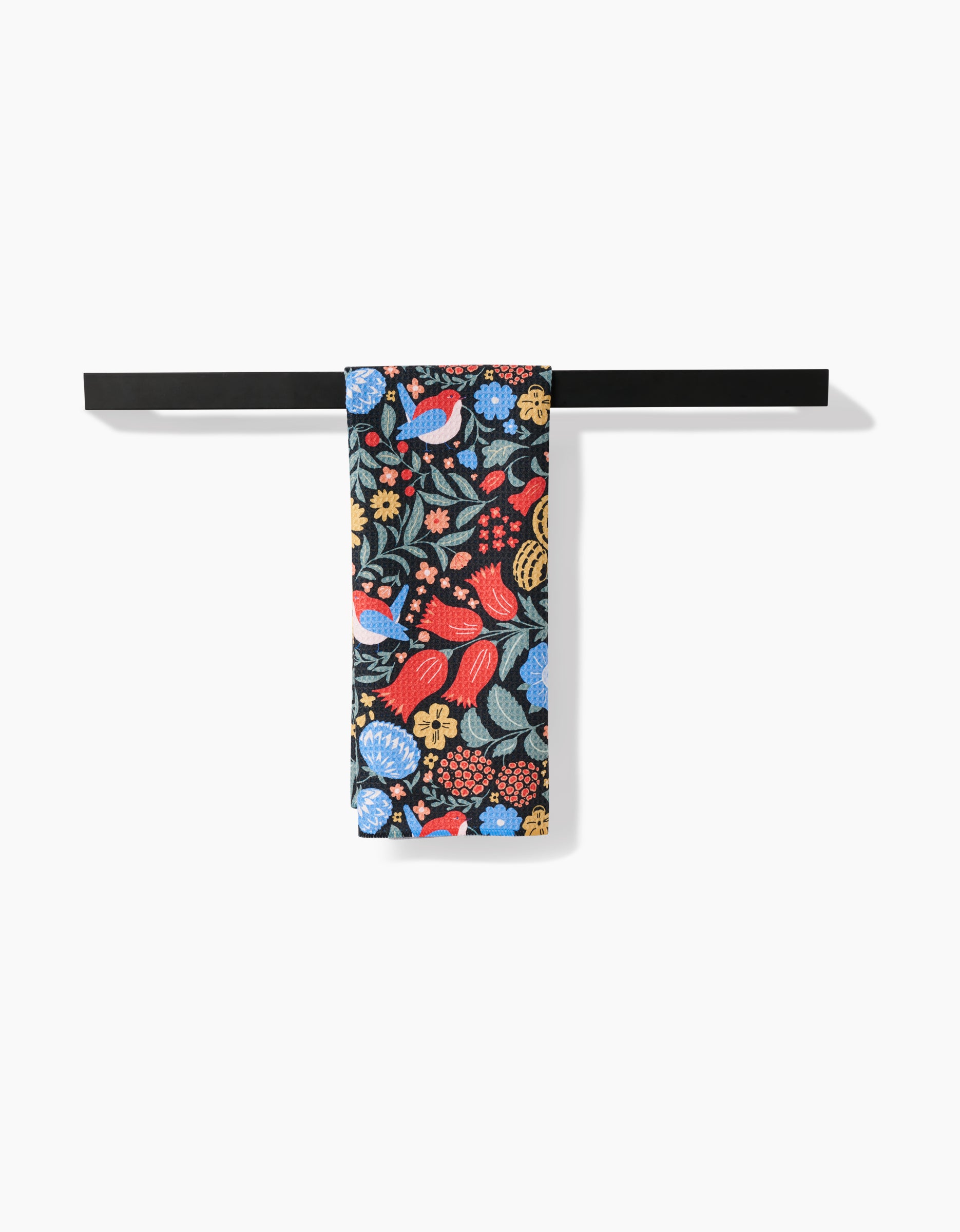A black towel bar displays the Danelys Sidron Marsh Wren tea towel, featuring a vibrant floral and leaf pattern in red, blue, yellow, and green on black Geoweave fabric for both style and absorbency.