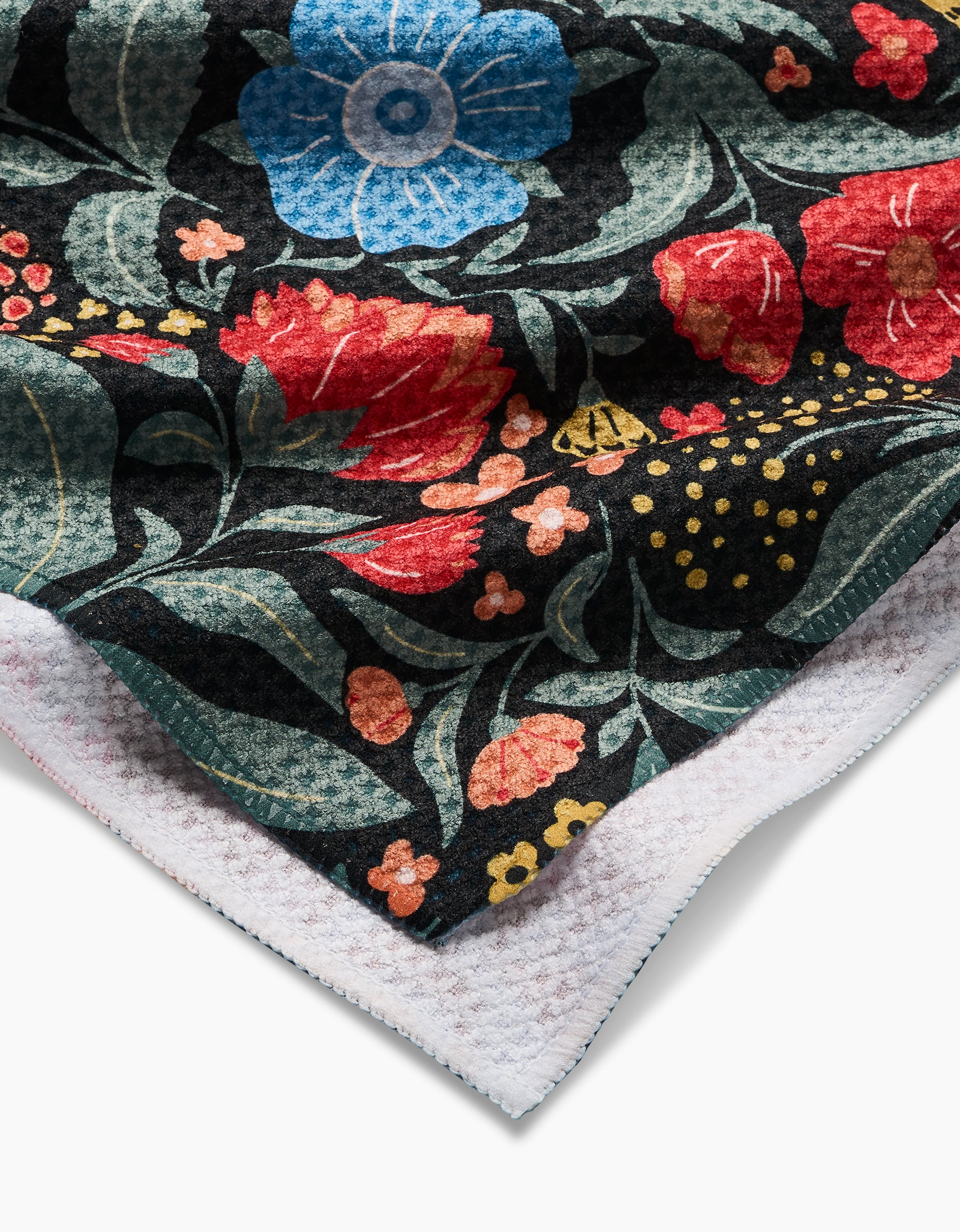 A close-up of the Marsh Wren tea towel by Danelys Sidron shows its colorful floral Geoweave fabric, with blue, red, and yellow flowers on black and green leaves, plus the ultra-absorbent white underside visible at the corner.