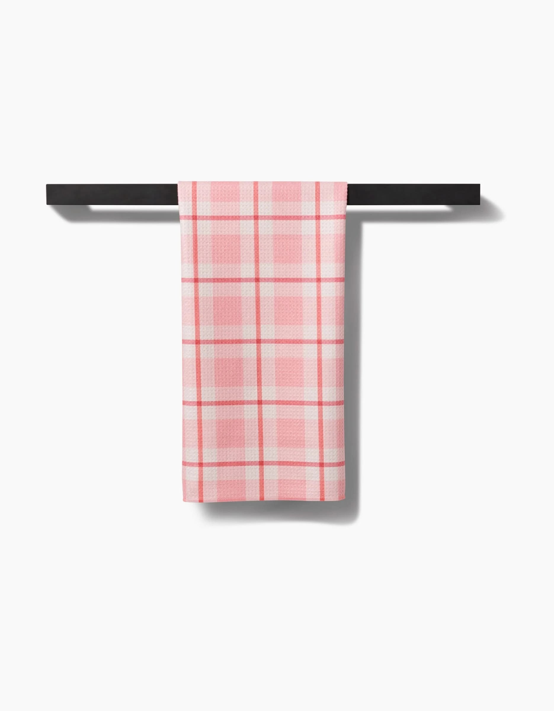 The Marva Lu tea towel by Kelsi Fullmer, featuring pink and white plaid Geoweave Fabric, is neatly folded and displayed on a modern black towel rack against a plain white background.