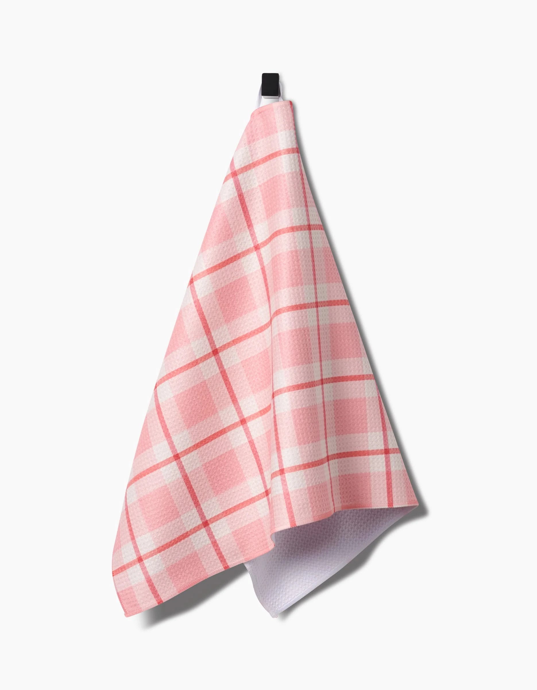The Marva Lu tea towel by Kelsi Fullmer, featuring a pink and white checkered pattern and made from recycled materials, hangs from a black hook against a white background.