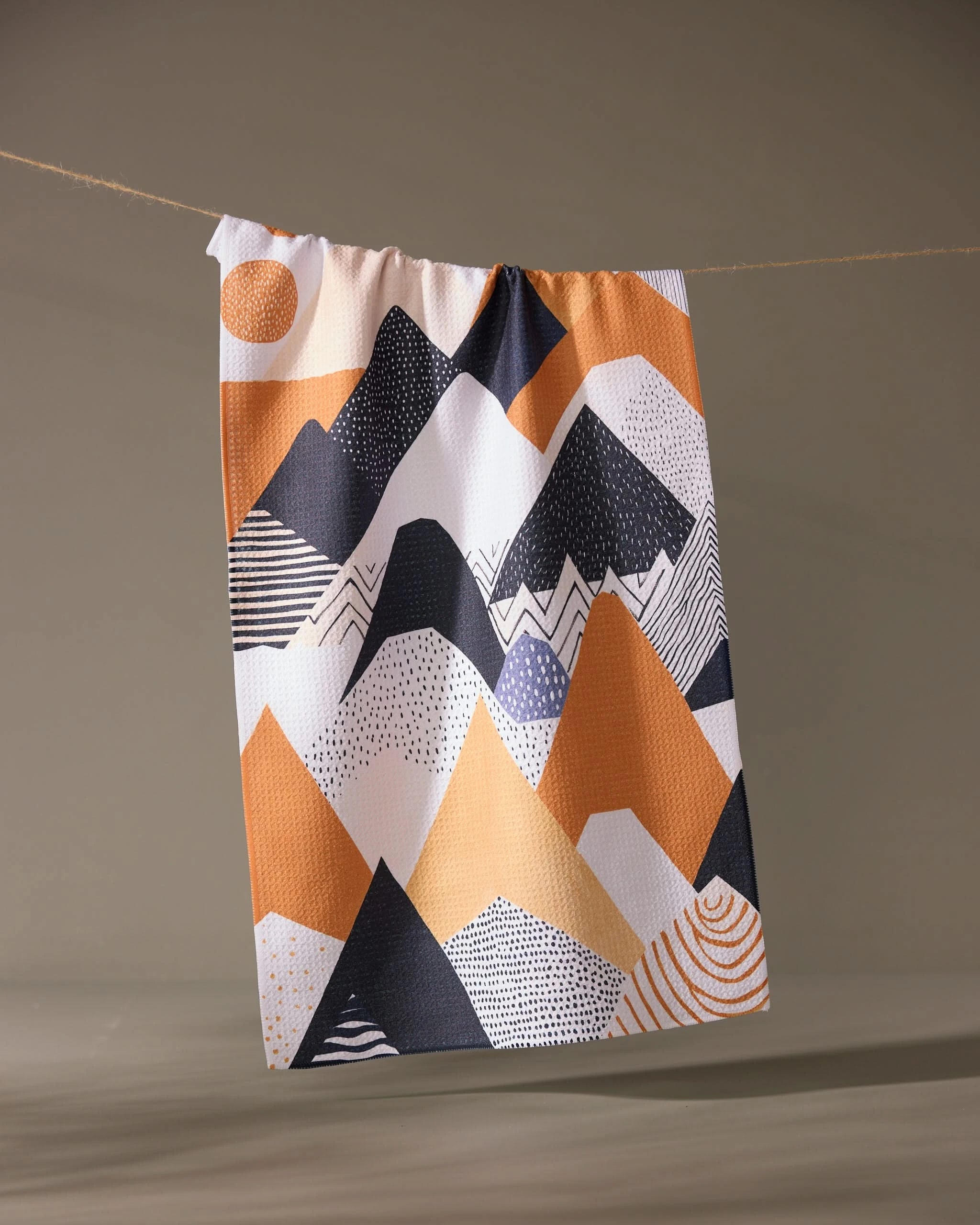 The Mesa Rise tea towel by Geometry hangs on a clothesline, showing off its vibrant abstract mountain design in orange, black, and white. Made from recycled materials, its an eco-friendly and stylish choice for your home.