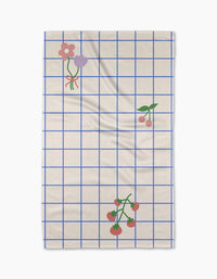 The Mini Harvest tea towel by Letters By Shells features a cream base with a blue grid, decorated with pink and purple flowers, cherries, and tomatoes—crafted from recycled materials for an eco-friendly touch.