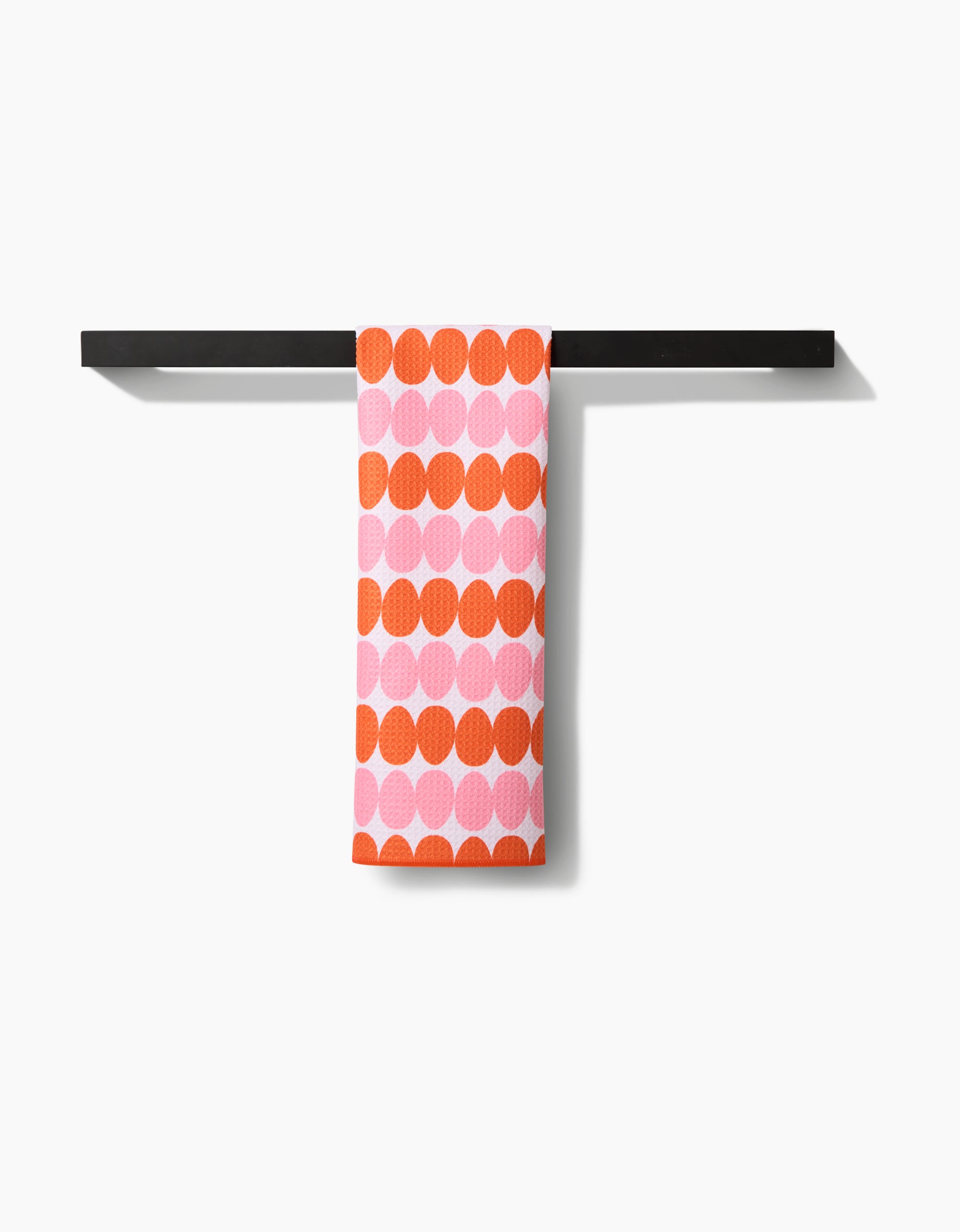 The Kayla Ann Modd Egg tea towel, featuring orange and pink circles, hangs neatly on a black towel bar against a white background. It is crafted from Geoweave fabric.
