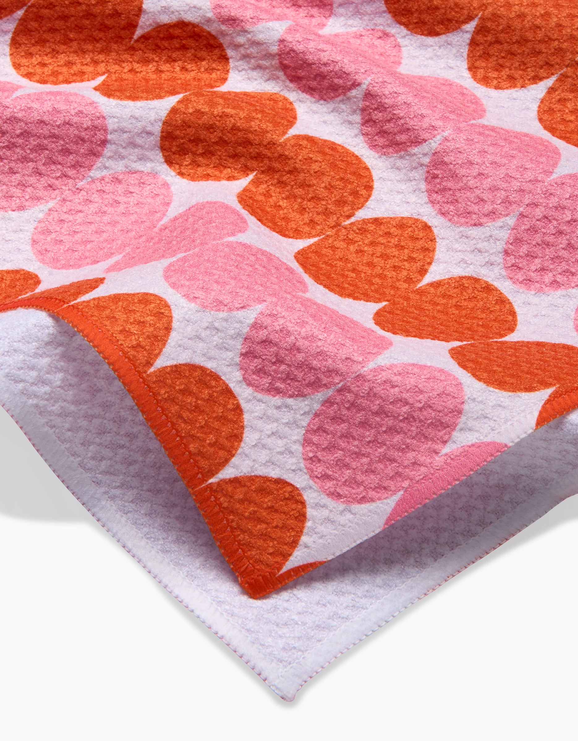A close-up of the Kayla Ann Modd Egg tea towel, showcasing overlapping pink and orange hearts on white Geoweave fabric with a folded corner displaying the reverse side.