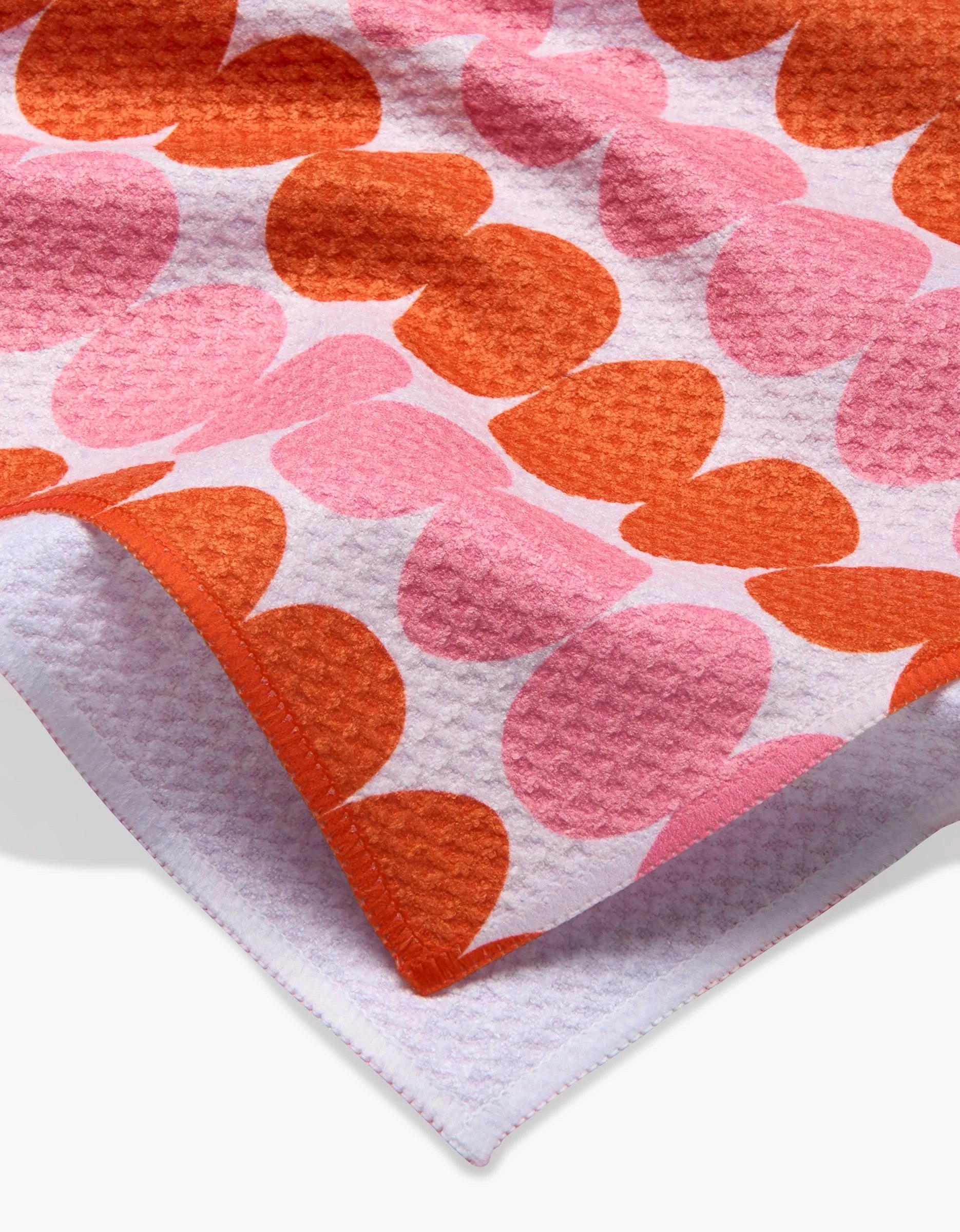 A close-up of the Kayla Ann Modd Egg tea towel, showcasing overlapping pink and orange hearts on white Geoweave fabric with a folded corner displaying the reverse side.