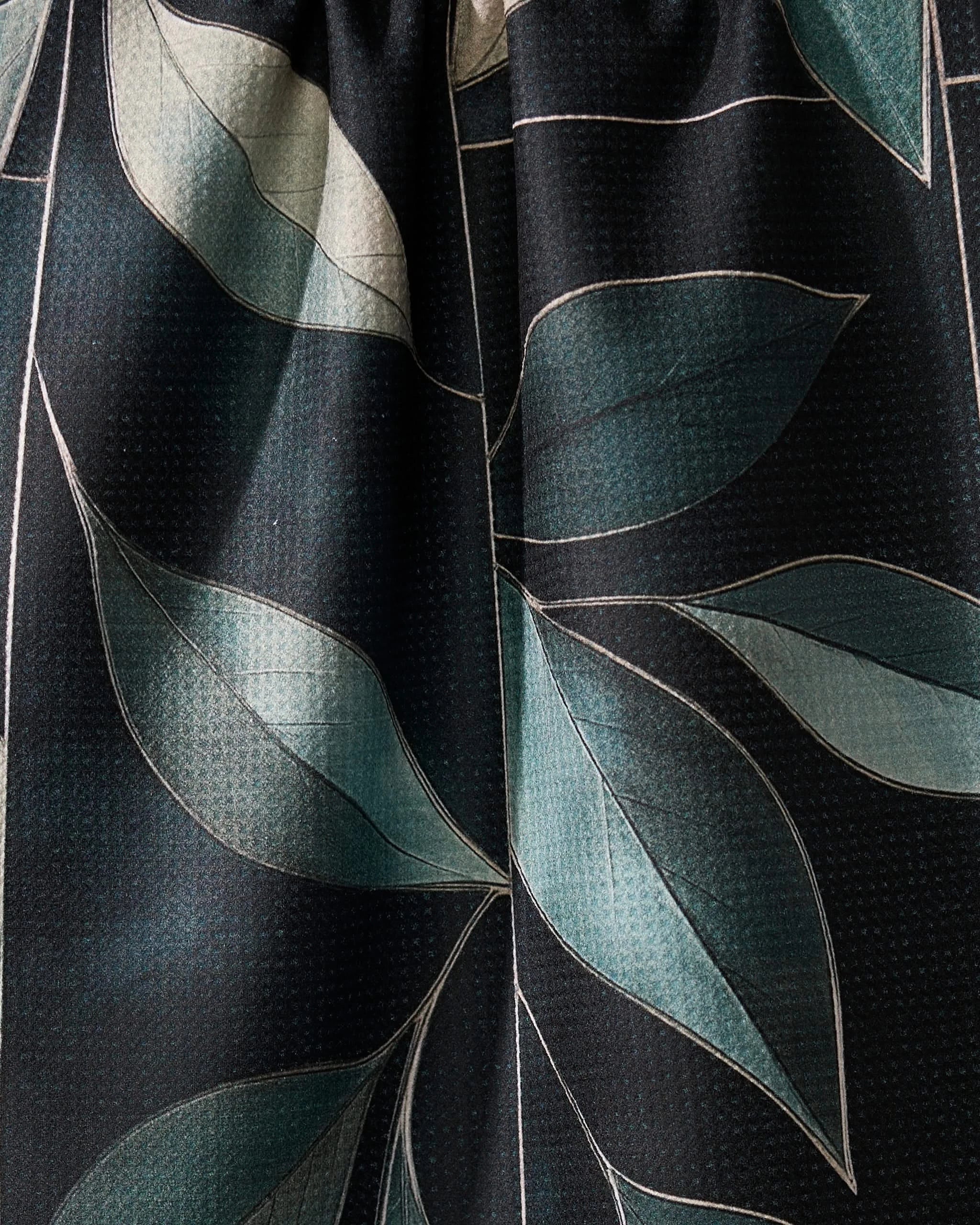 A close-up of Modern Organic by Geometry shows dark fabric with large teal and light green leaves outlined in white. This recycled kitchen towel has a subtle textured surface, combining bold, modern botanical design with sustainability.