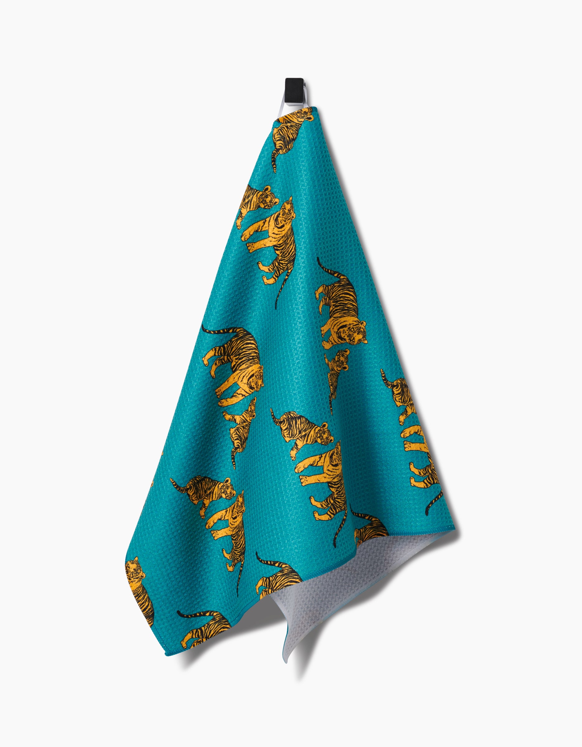 The Mommy and Me tea towel by Sarah Holden, made from recycled materials, displays a turquoise design with illustrated orange tigers and hangs from a black hook against a white background.