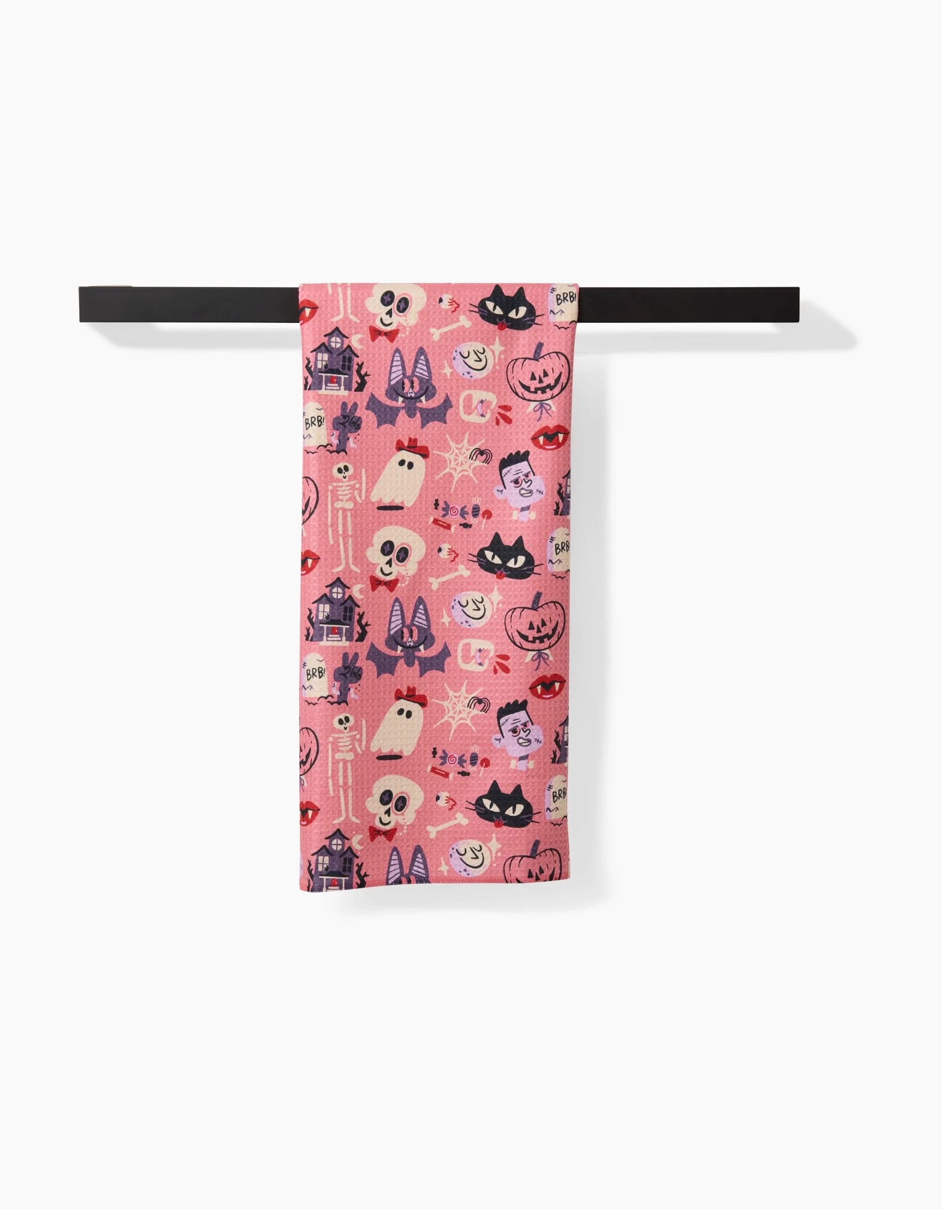 The Carly Berry Monstermash tea towel, in pink, hangs on a black rod and features a Halloween pattern with ghosts, pumpkins, bats, black cats, haunted houses, and stars.