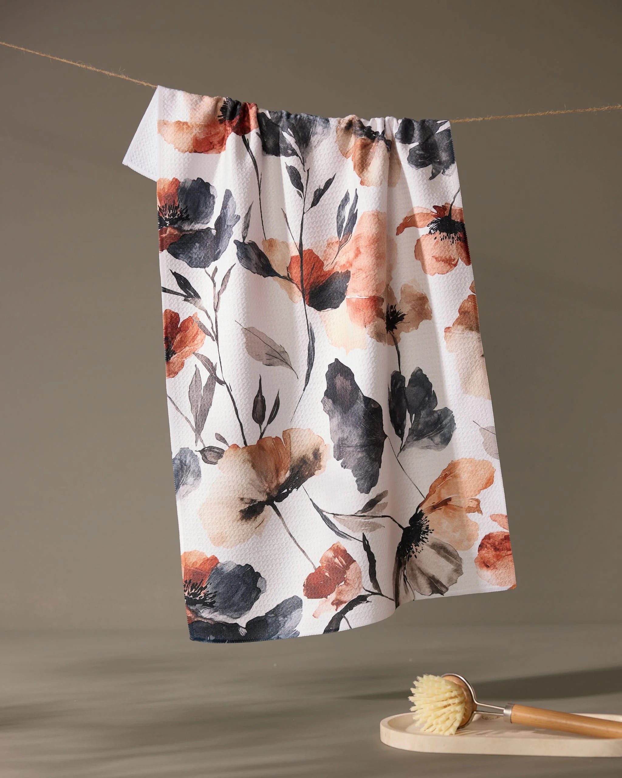 The Moody Poppy tea towel by Geometry, featuring orange, black, and gray floral designs on recycled fabric, hangs on a string above a wooden dish brush set on a small round surface against a plain neutral background.