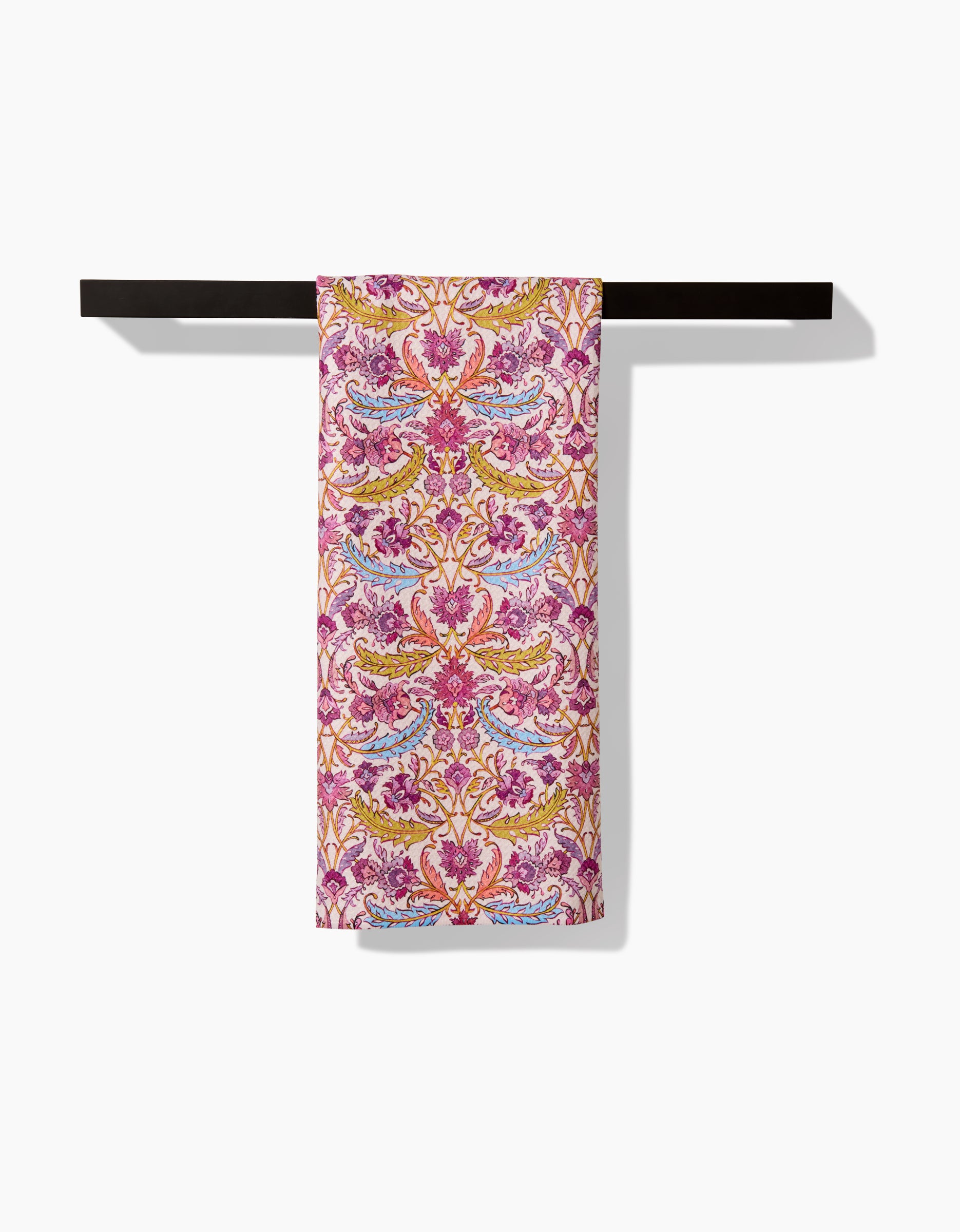 The Morning Glory tea towel by Maayan Alper-Swan features an ornate, symmetrical floral design in pink, gold, and light blue, neatly draped over a sleek black towel bar on a white background.