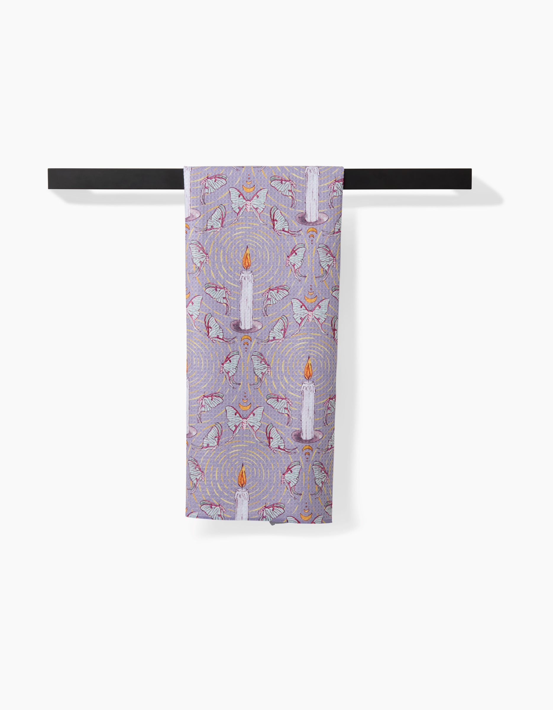 The Moth To A Flame tea towel by Maayan Alper-Swan, made from recycled materials and featuring white candle illustrations, hangs over a black rod against a plain white background.