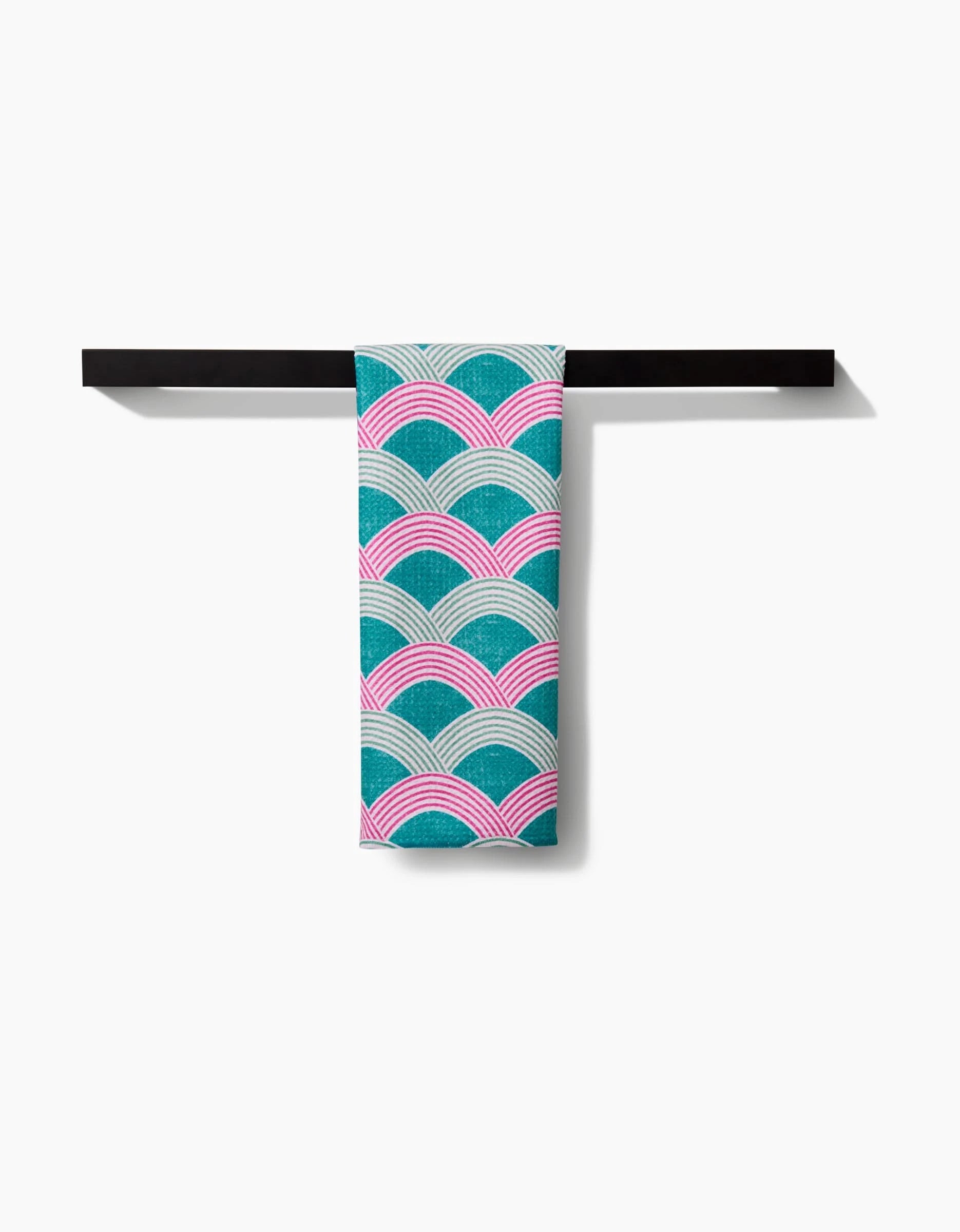 The Nalin tea towel by Amy MacCready, featuring a teal, pink, and white wave pattern on soft Geoweave fabric, hangs on a sleek black towel bar against a white background.