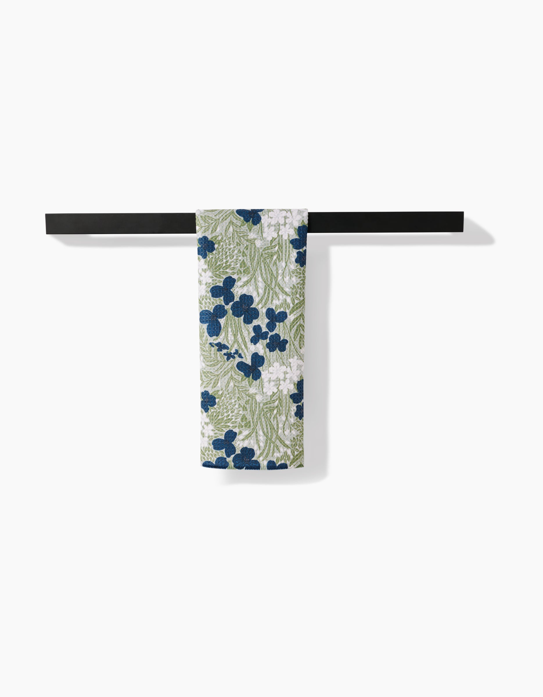 The Navy And Sage Meadow towel by Katie OShea, featuring blue flowers and green leaves on ultra-absorbent Geoweave fabric, hangs on a minimalist black rack against a white background.