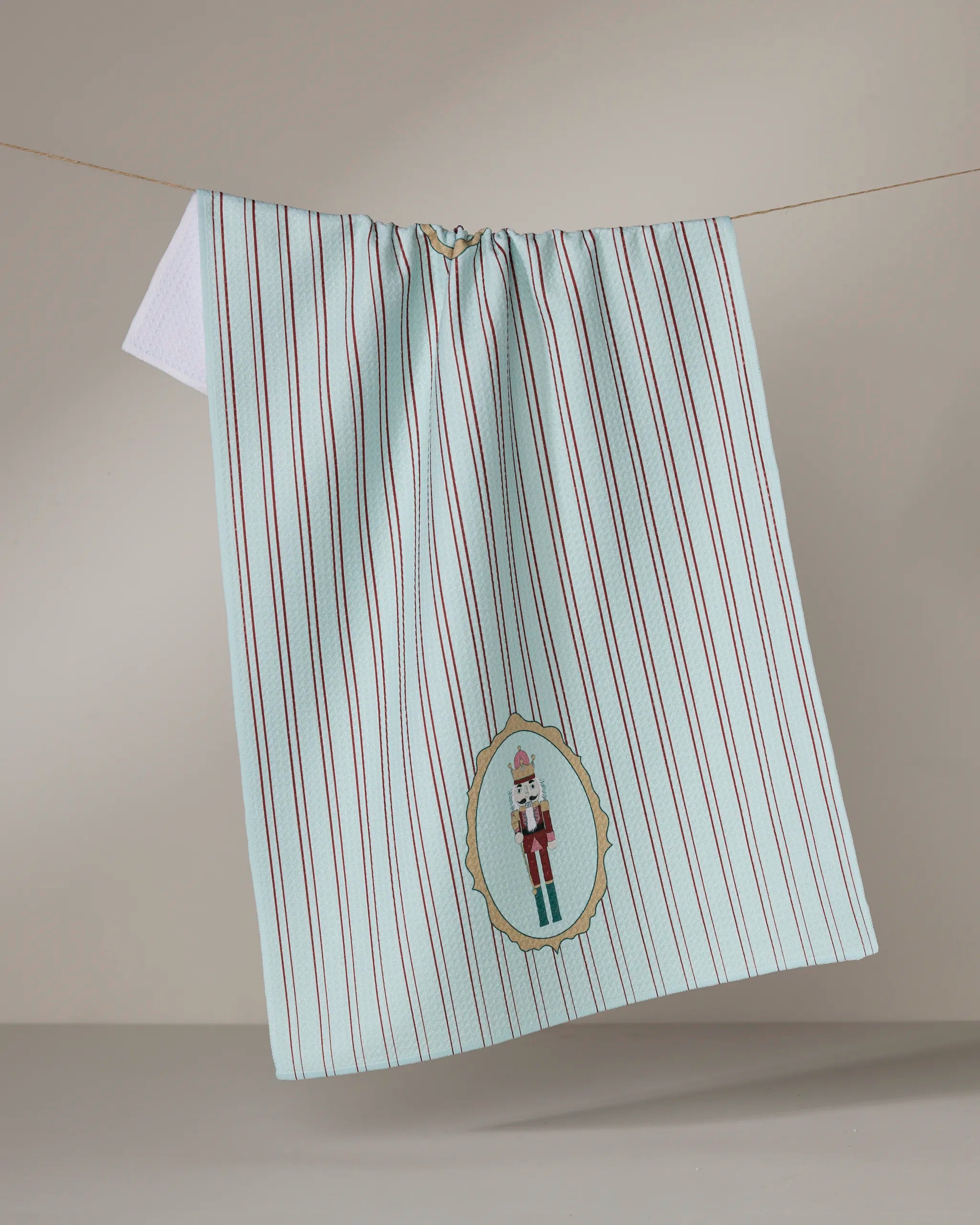 The Nutcracker Intermission by Geometry is a blue and white striped tea towel with thin red lines, ultra-absorbent, and features an embroidered nutcracker design in a decorative oval near the bottom edge.