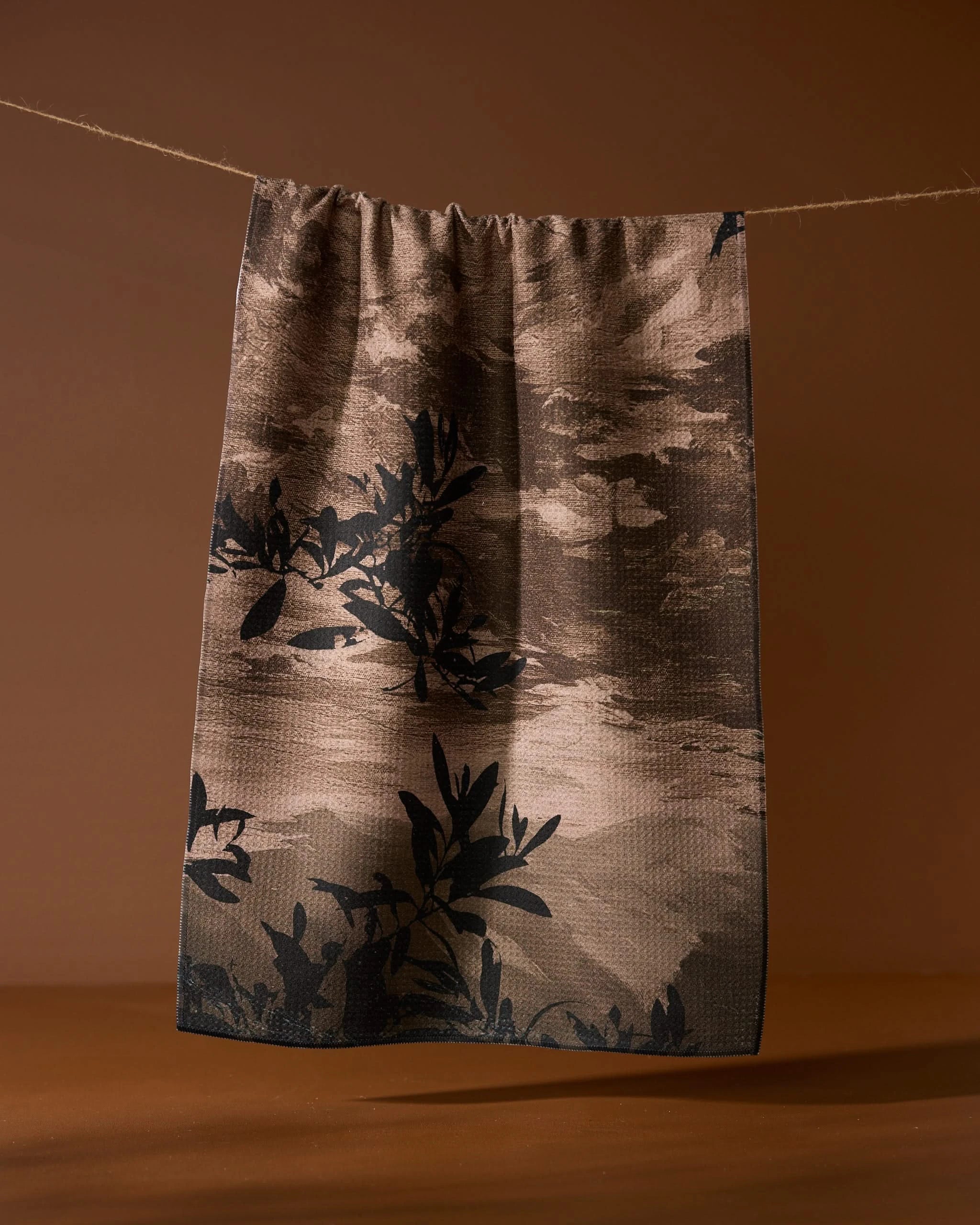 The Geometry Oaklen tea towel, featuring a dark leafy branch pattern in brown and beige and made from recycled materials, hangs from a string against a solid brown background.