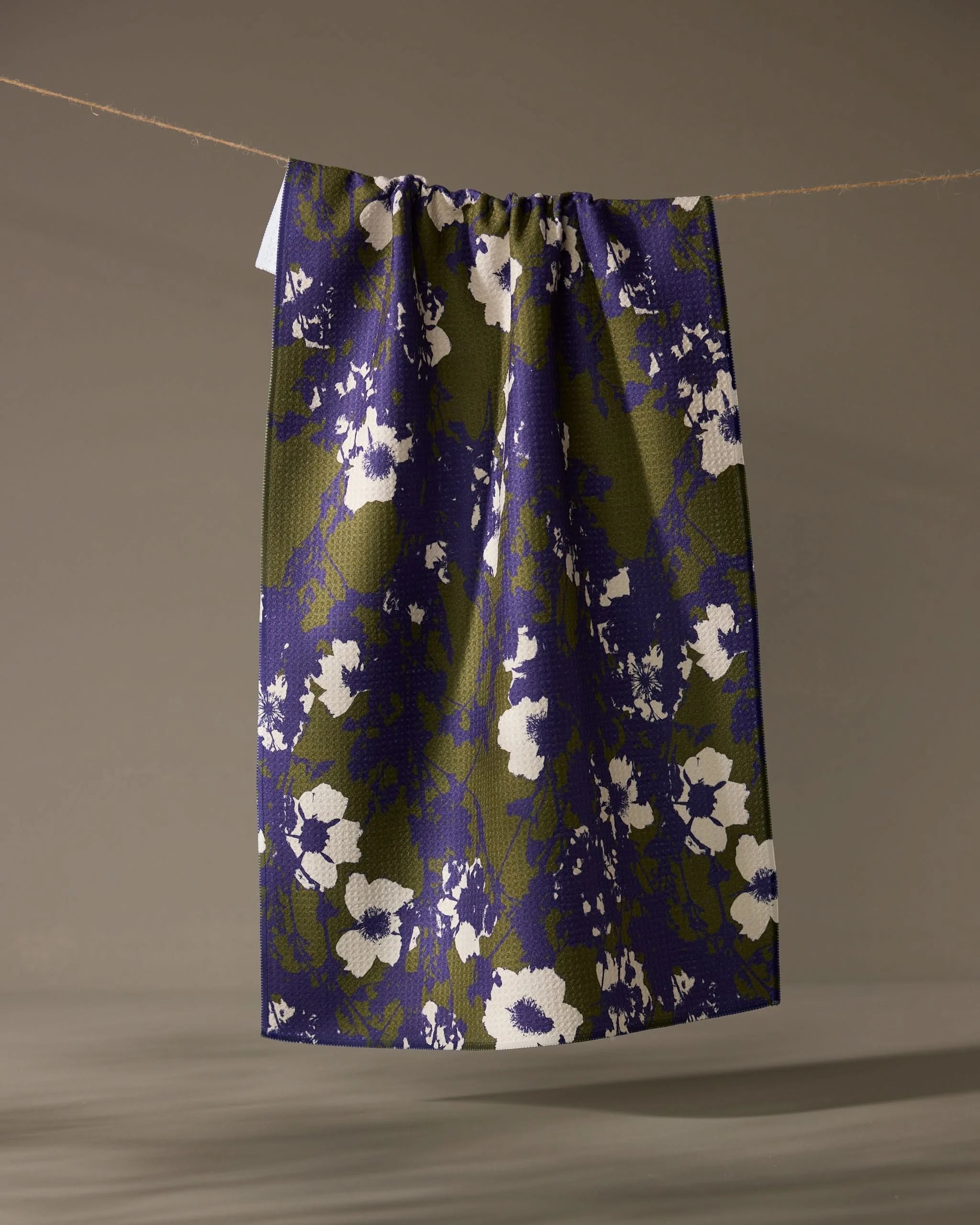 The Oliveberry skirt by Geometry, knee-length with a gathered waistband, made from quick-drying recycled materials and adorned with a blue and olive green abstract floral print, hangs from a clothesline against a neutral background.