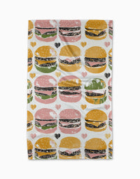 The Geometry On The Grill Tea Towel features a repeating pattern of stylized hamburgers in yellow, pink, and green buns with small hearts on white Geoweave fabric made from recycled materials.