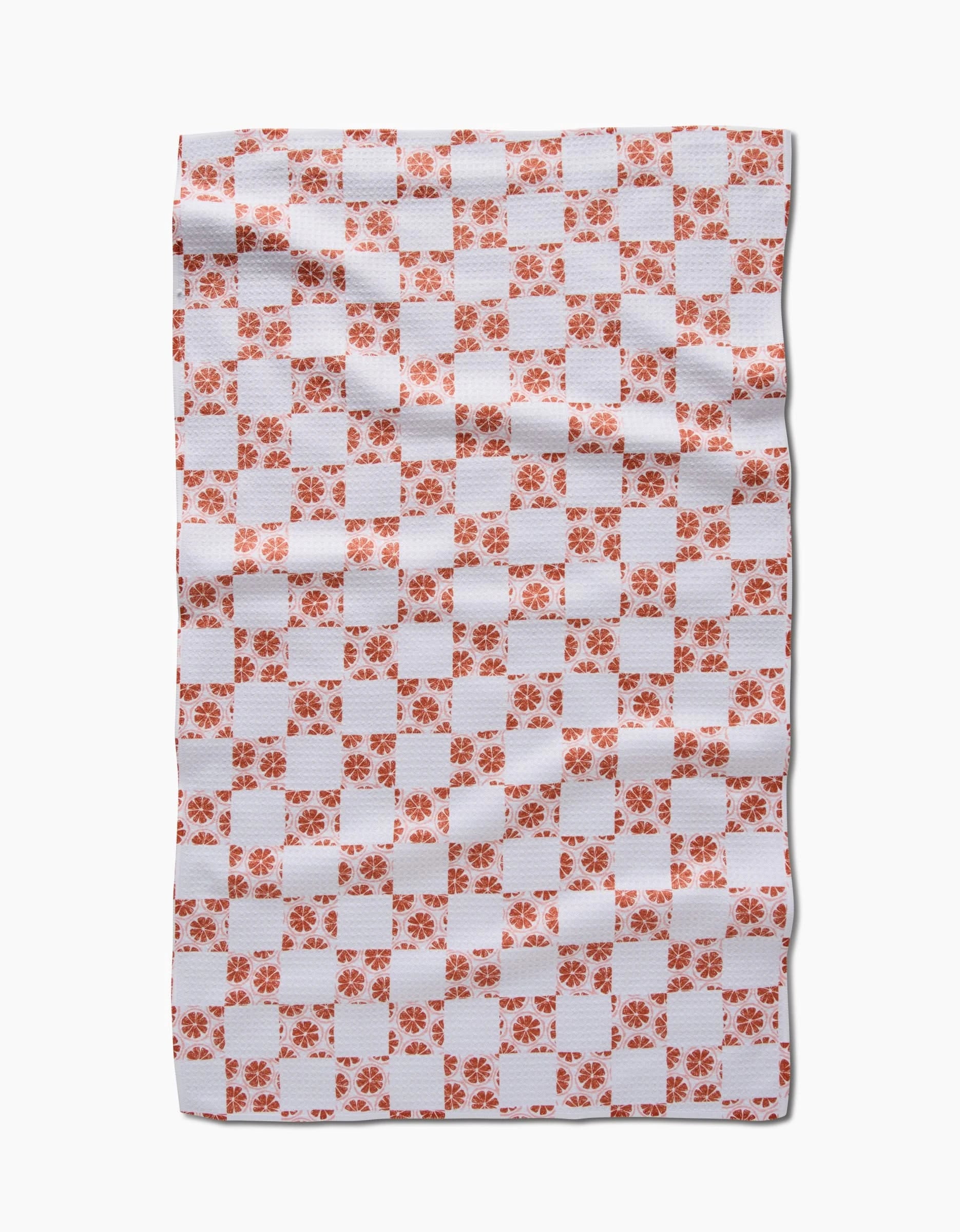 The Orange Checker by Teri the Butter is a rectangular white tea towel with an orange checkered pattern and floral designs, crafted from Geoweave™ fabric. Cloth appears slightly wrinkled, shown on a plain white background.