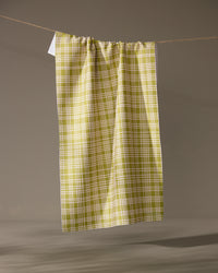 The Orchard Plaid Green tea towel by Geometry hangs vertically from a string against a gray background. Made from recycled materials, its lightweight fabric is gathered at the top for an eco-friendly, stylish touch.