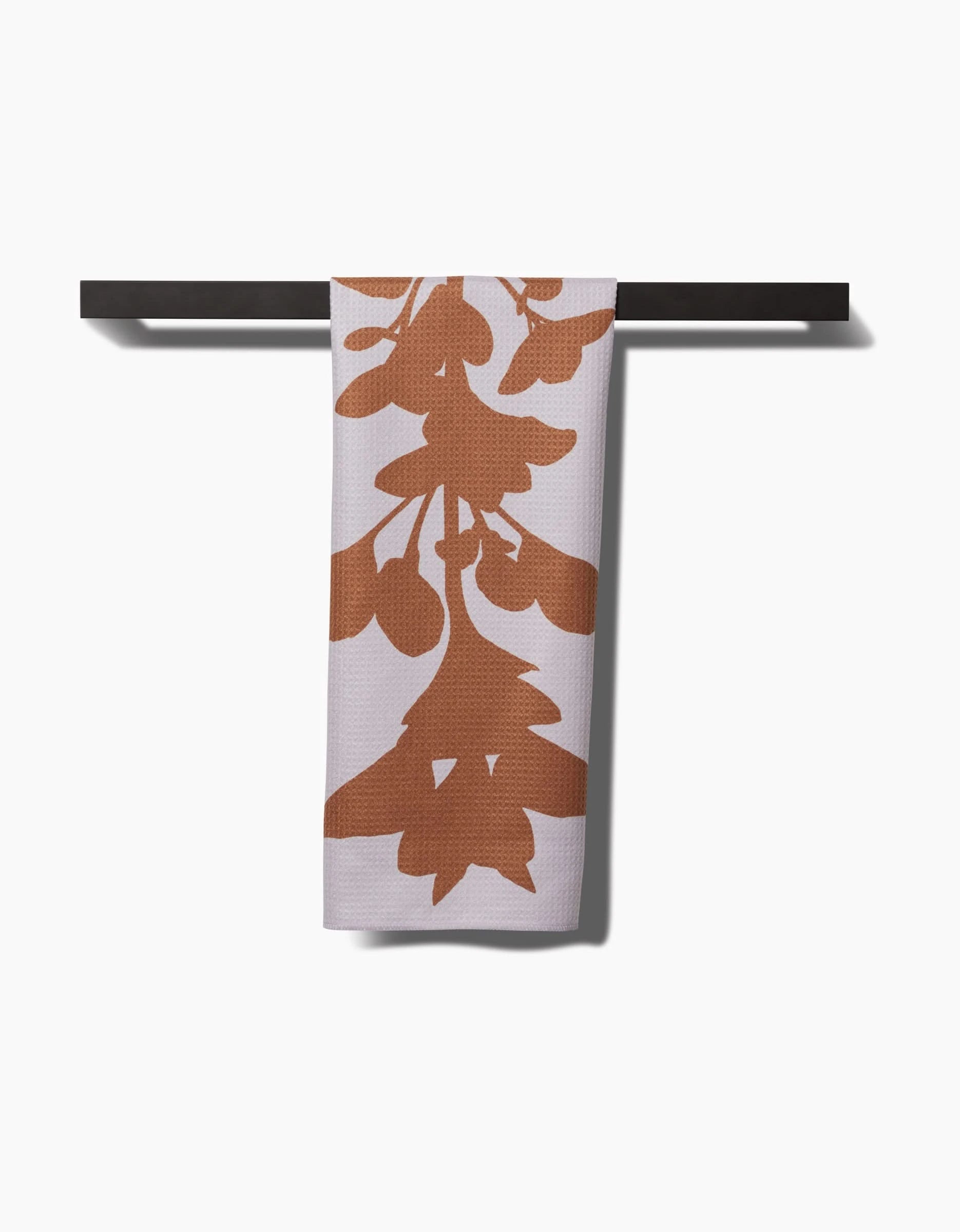 The PrintedPlant Oregano tea towel, made from durable Geoweave™ fabric, features a bold rust-colored leaf pattern and hangs neatly on a dark towel bar against a white background.