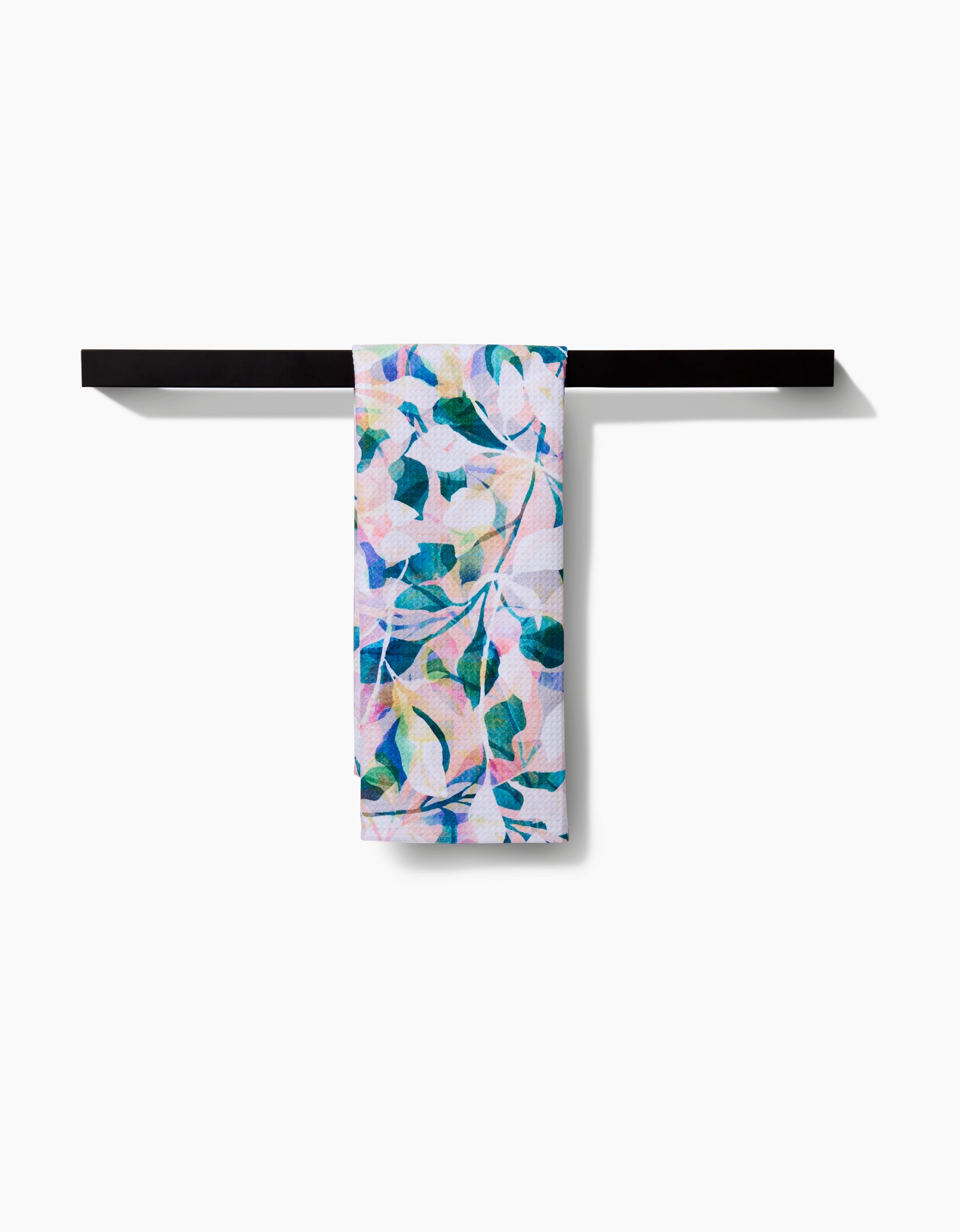 The Painted Foliage tea towel by Rachel Parker, featuring a vibrant green, blue, and pink leaf pattern, hangs over a sleek black bar. Made from recycled materials and set against a white background.