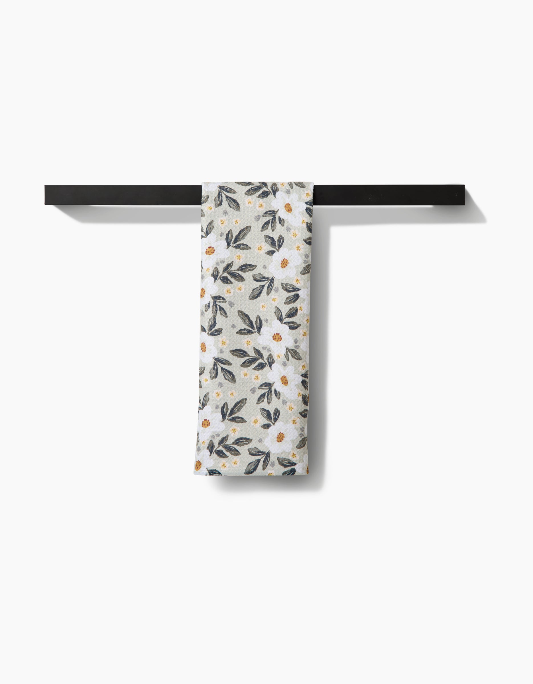 The Painted Spring Flowers tea towel by Teri the Butter, crafted from soft Geoweave™ fabric, features a floral design with white flowers and green leaves and hangs neatly on a dark horizontal bar against a white background.