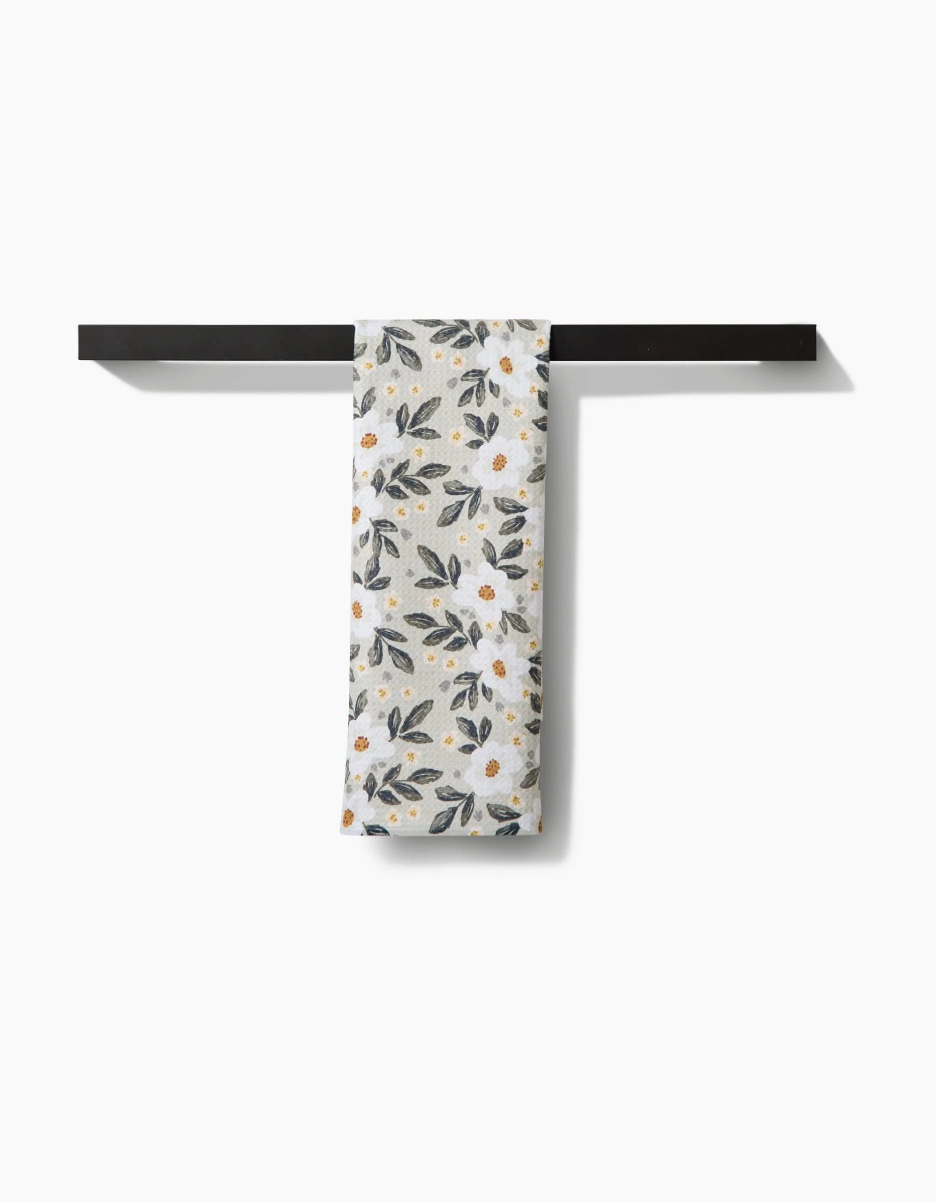 The Painted Spring Flowers tea towel by Teri the Butter, crafted from soft Geoweave™ fabric, features a floral design with white flowers and green leaves and hangs neatly on a dark horizontal bar against a white background.