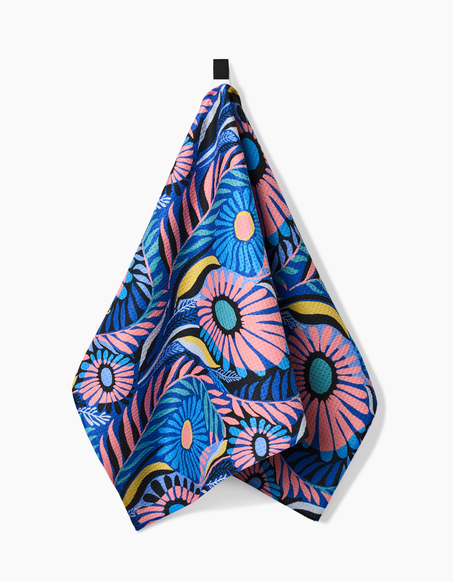 The Palm Beach tea towel by Masha Volnova features a vibrant blue, pink, yellow, and black floral pattern on recycled material, hanging from a black loop against a plain white background.