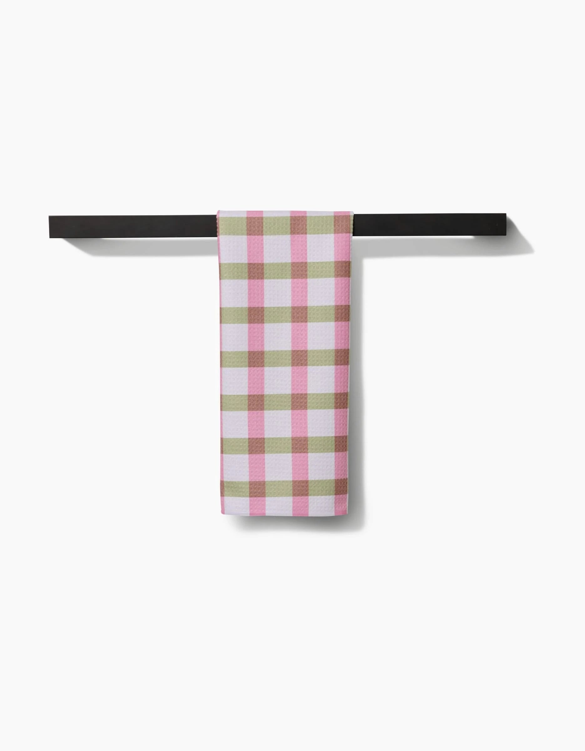 The Patricia Plaid Tea Towel by Laura Prietto, featuring a pink, white, and beige check pattern, hangs neatly folded on a black bar and is made from Geoweave™ fabric for softness and durability.