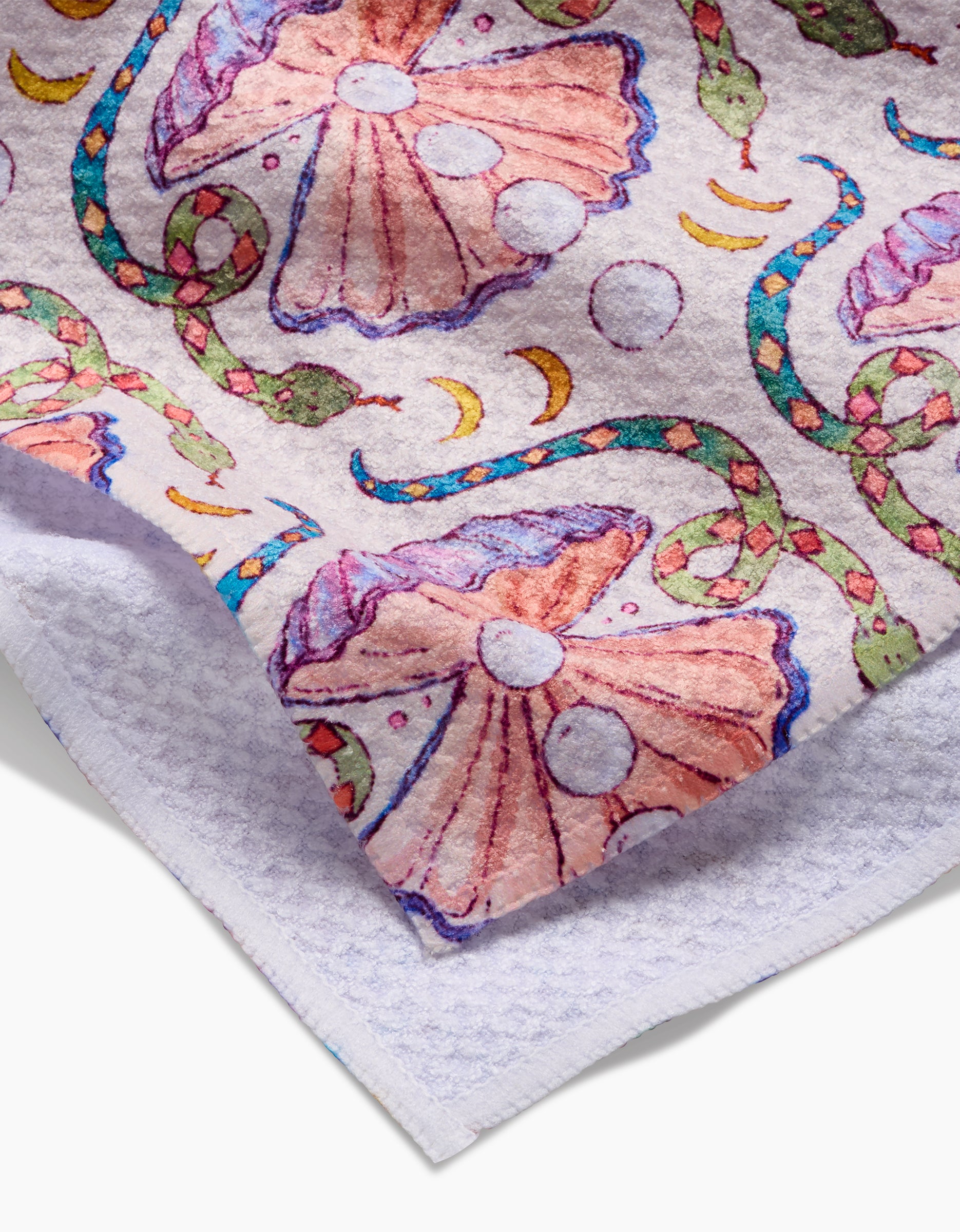 A close-up of the Pearls Of The Moon towel by Maayan Alper-Swan, with pink seashells, snakes, moons, and bubbles on textured white Geoweave fabric. Its folded corner shows off the ultra-absorbent plush thickness.