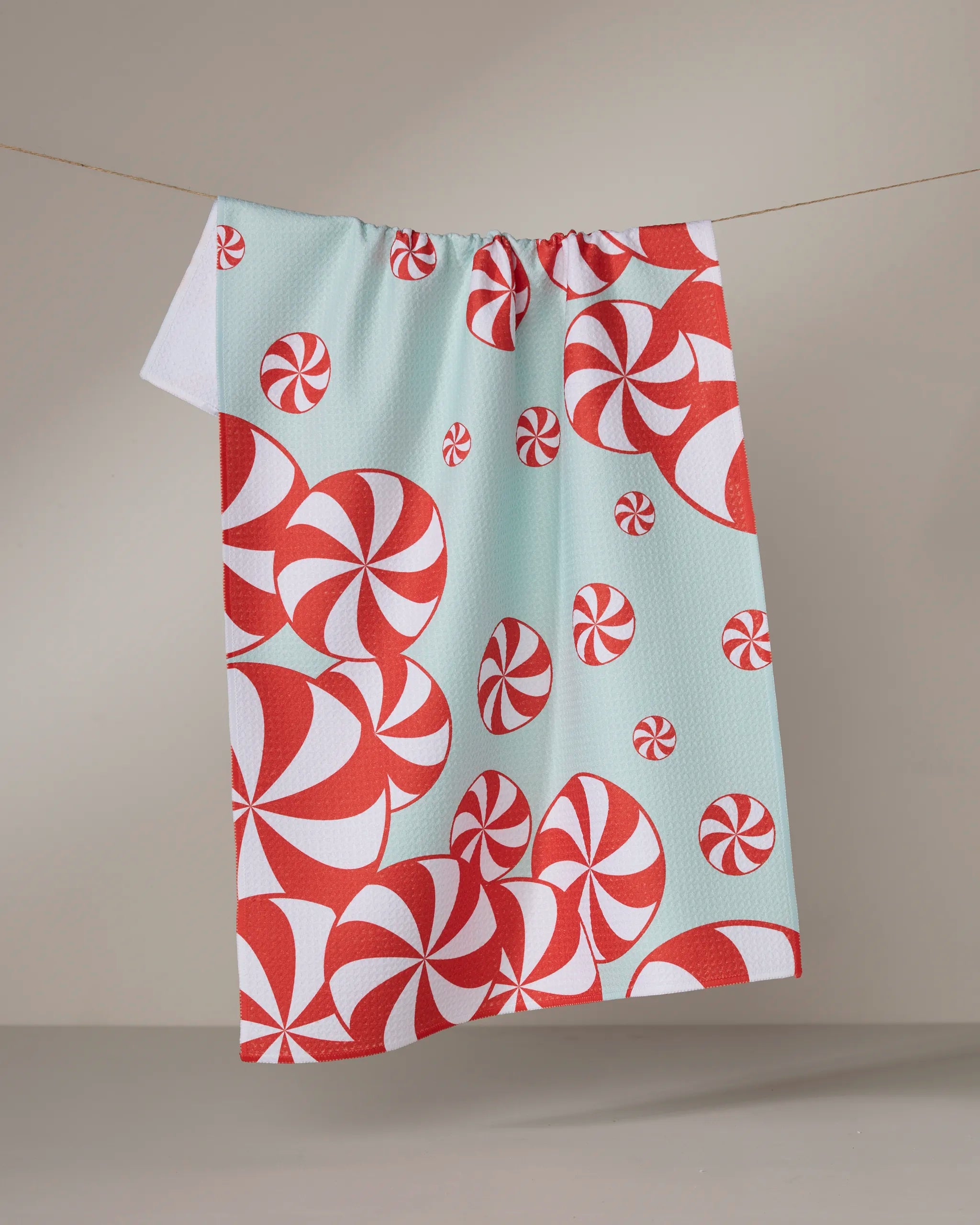 The Geometry Peppermint Swirl tea towel, decorated with red and white peppermint candies on a light blue background, hangs on a clothesline against a neutral backdrop.