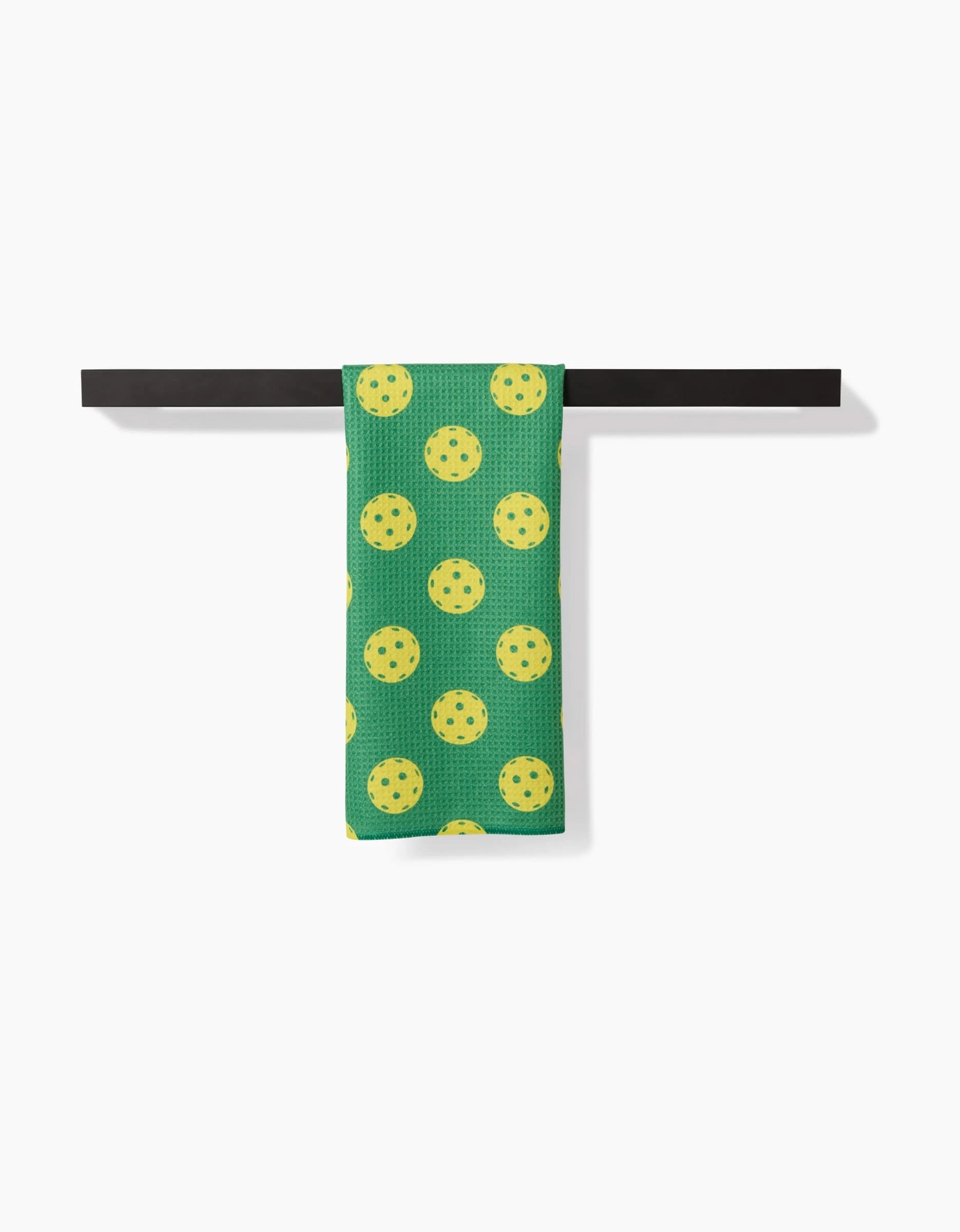 A GEOMETRY Pickleball tea towel with green fabric and yellow pickleball-like circles is neatly folded over a black towel bar against a white background.