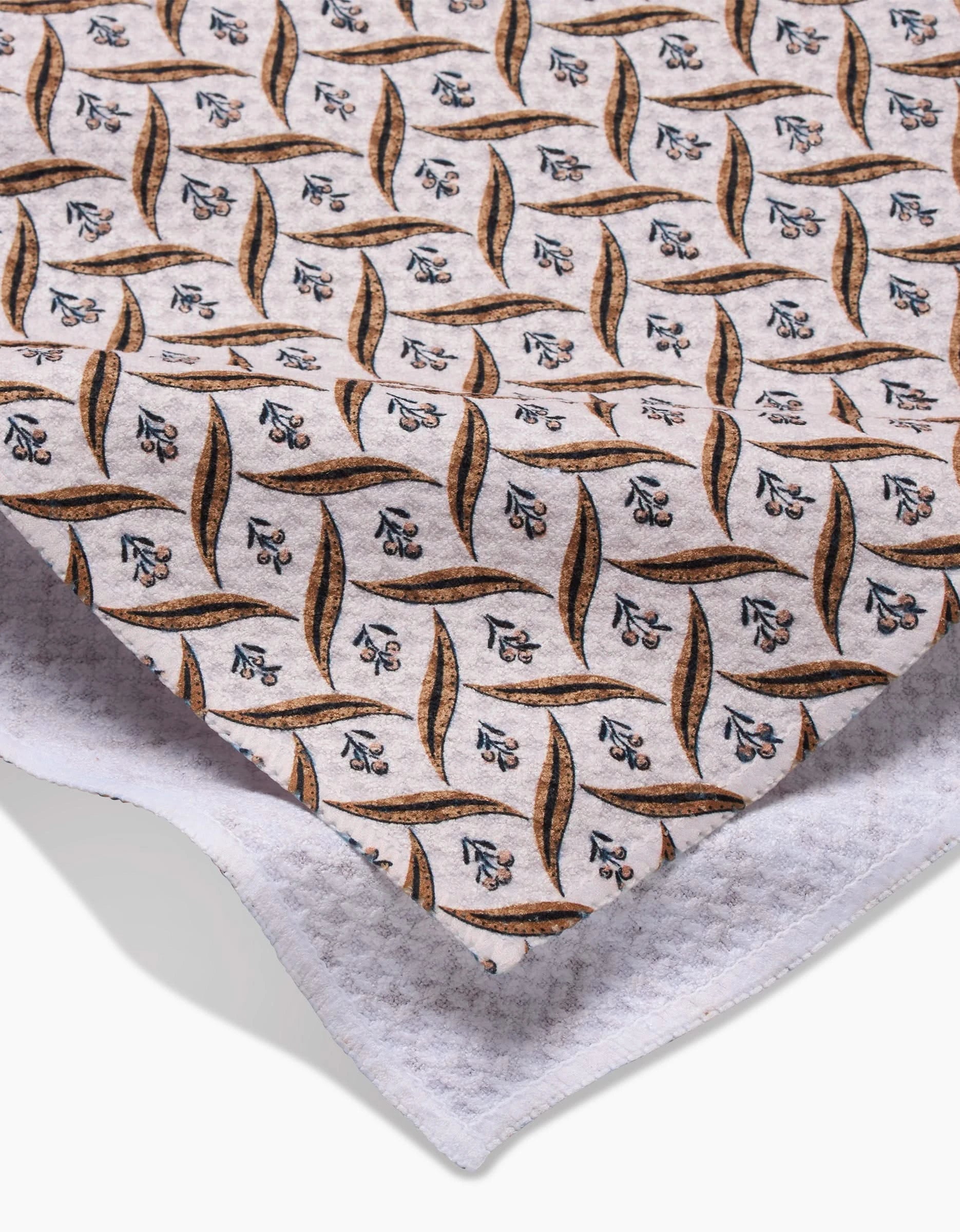 A close-up of Hufton Studios Plated Fall Berry Natural fabric, featuring a repeating leaf and floral design in brown, blue, and black on white, made from recycled materials and layered over textured white cloth.
