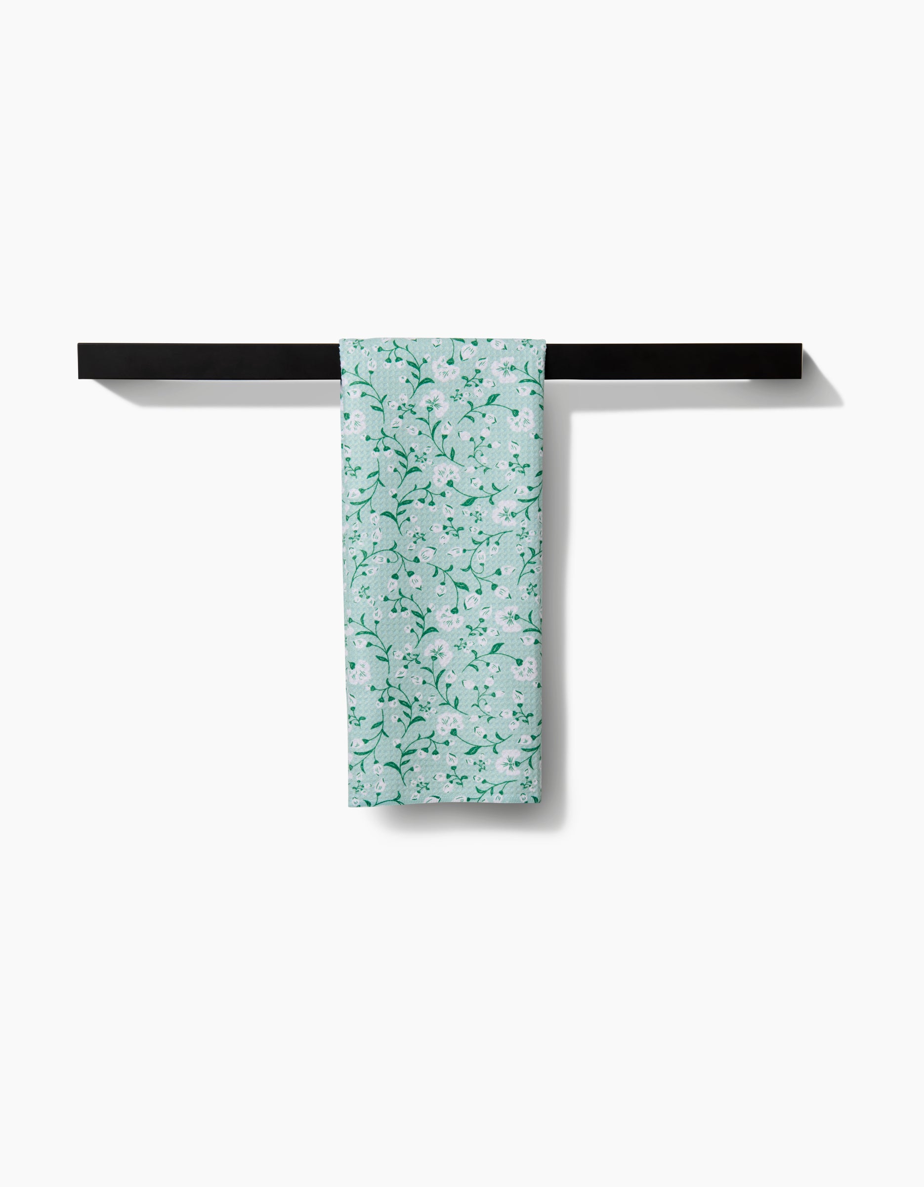 A Plumbago Bloom tea towel by Danelys Sidron, featuring a light green floral design and crafted from Geoweave fabric, hangs neatly folded over a simple black bar on a white wall.