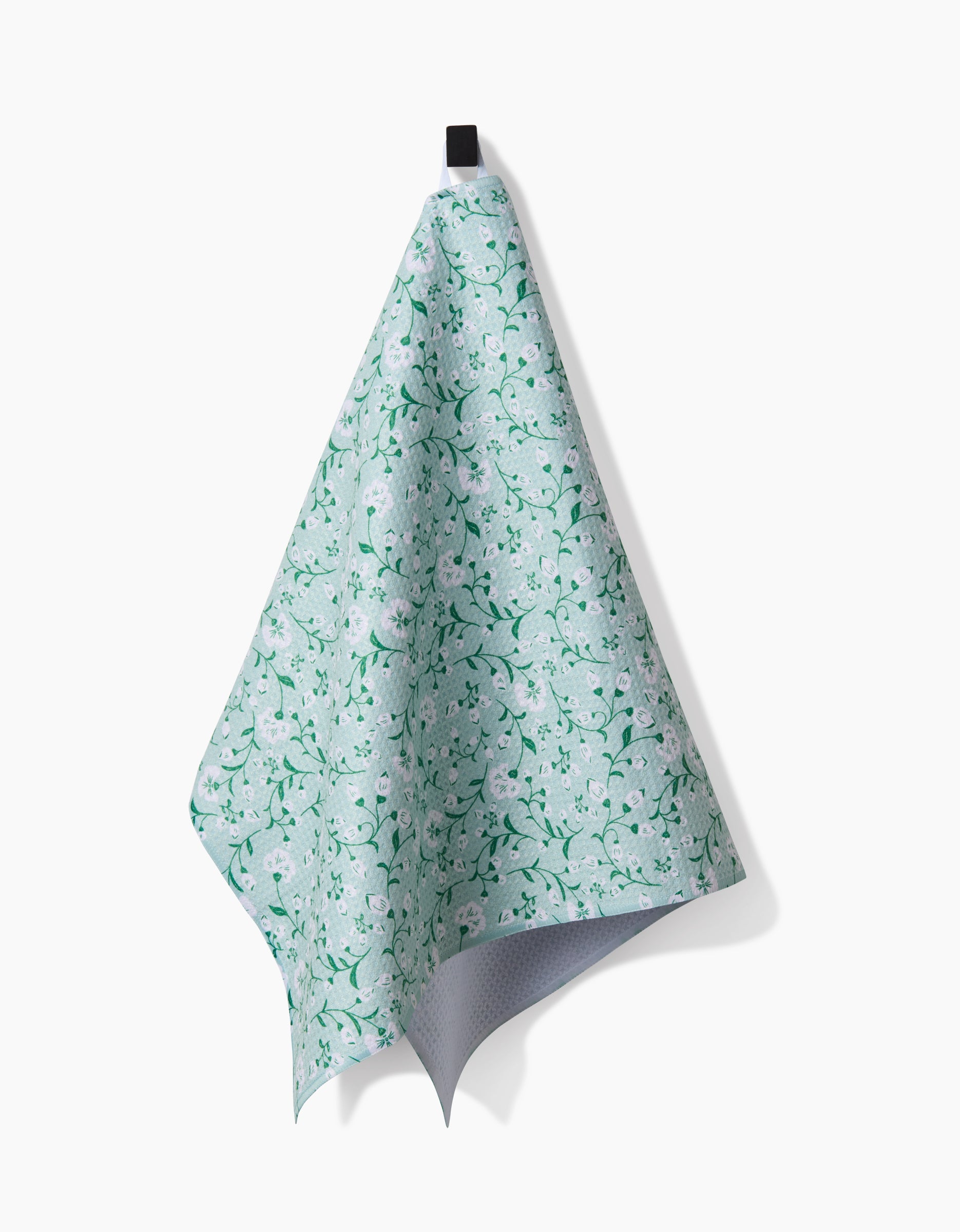 A green and white floral-patterned Plumbago Bloom tea towel by Danelys Sidron hangs neatly from a black hook against a plain white background.