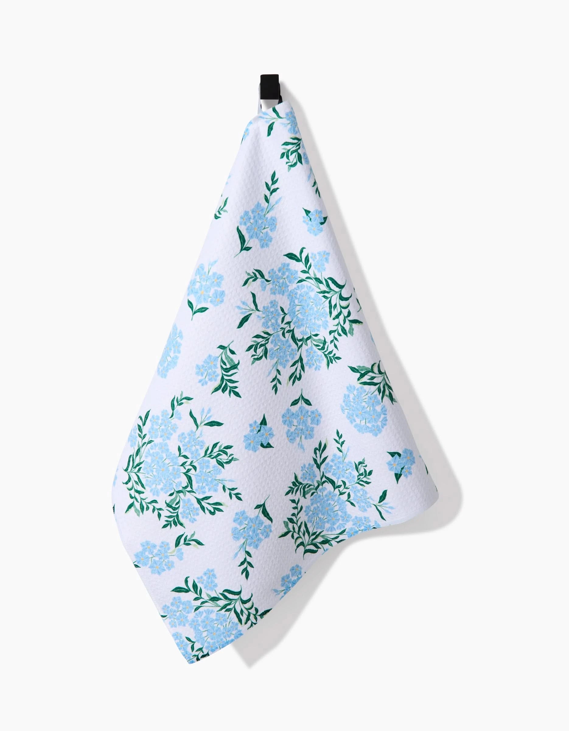 The Plumbago towel by Danelys Sidron, crafted from ultra-absorbent Geoweave™ fabric and adorned with blue floral and green leafy patterns, hangs from a black hook against a plain white background.