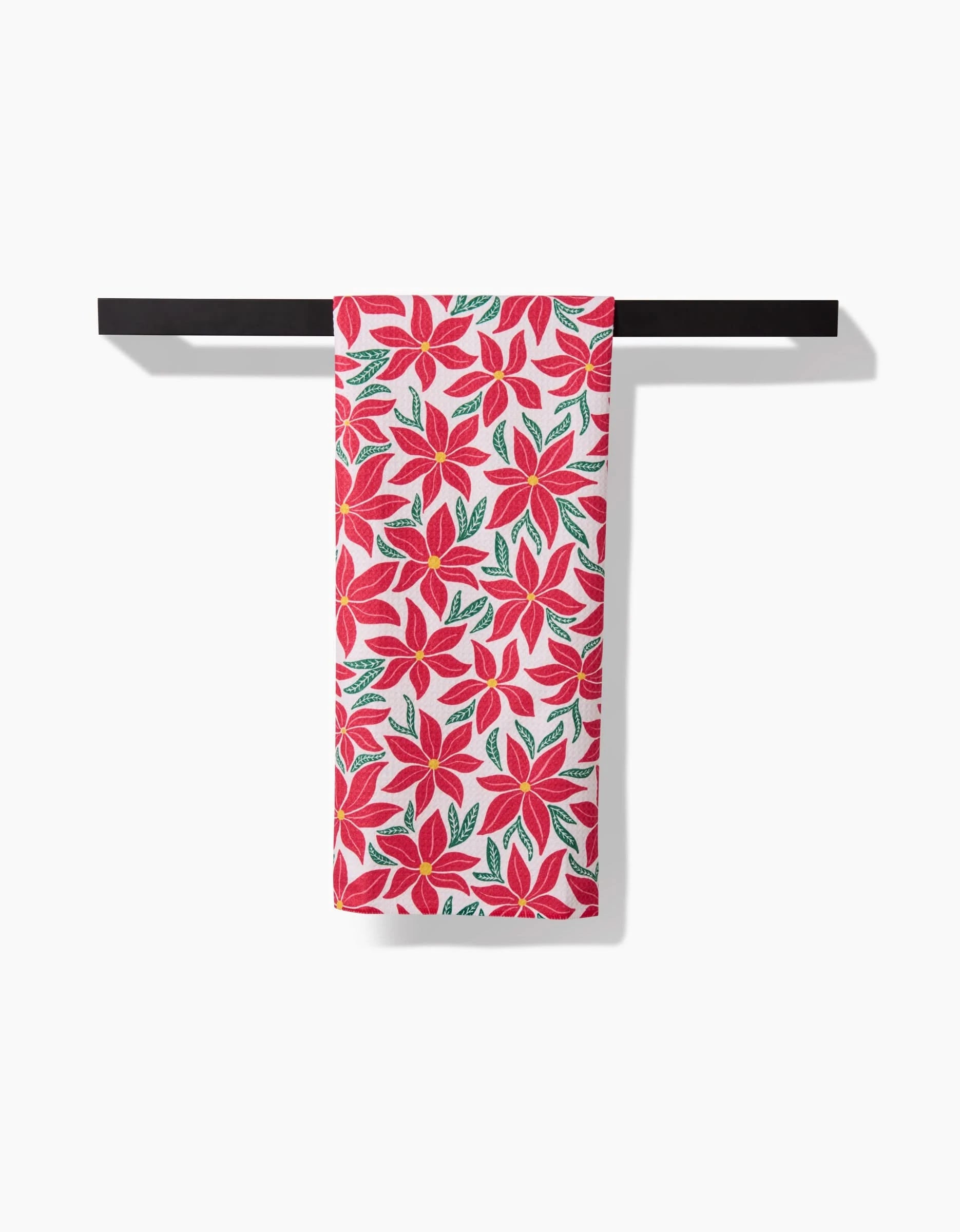 The Kayla Ann Poinsettia Party kitchen tea towel, featuring a vibrant red poinsettia and green leaf design, is shown neatly folded over a black horizontal bar against a plain white background.