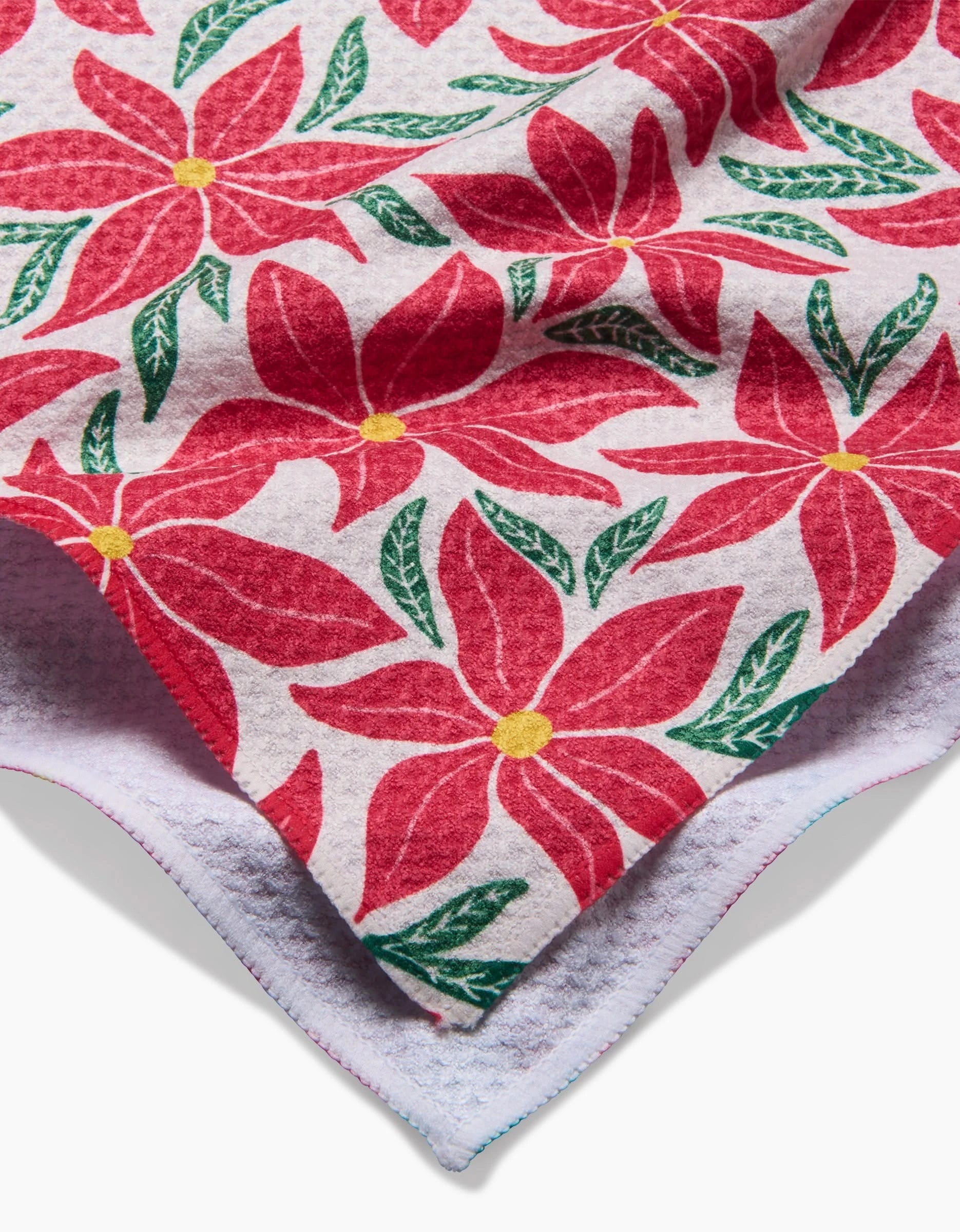 A close-up of the Kayla Ann Poinsettia Party kitchen tea towel, featuring a red poinsettia and green leaf pattern on white, with a folded corner showing visible stitching and its ultra-absorbent quality.