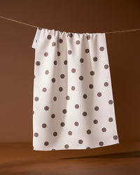 The Polka Picnic tea towel by Geometry, featuring black polka dots on white, hangs on a string against a brown background.