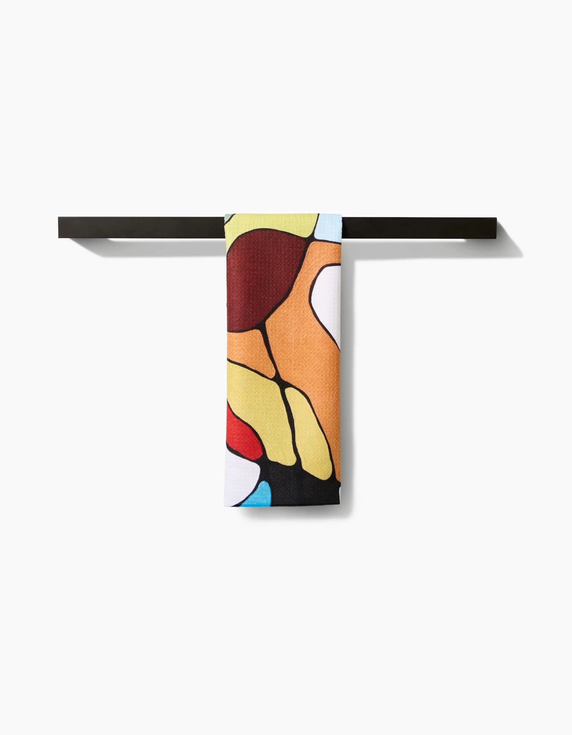 The Poppies on Blue tea towel by Rachel Austin, featuring an abstract pattern of red, yellow, orange, black, and blue shapes on Geoweave fabric, hangs neatly over a simple black rack against a white background.