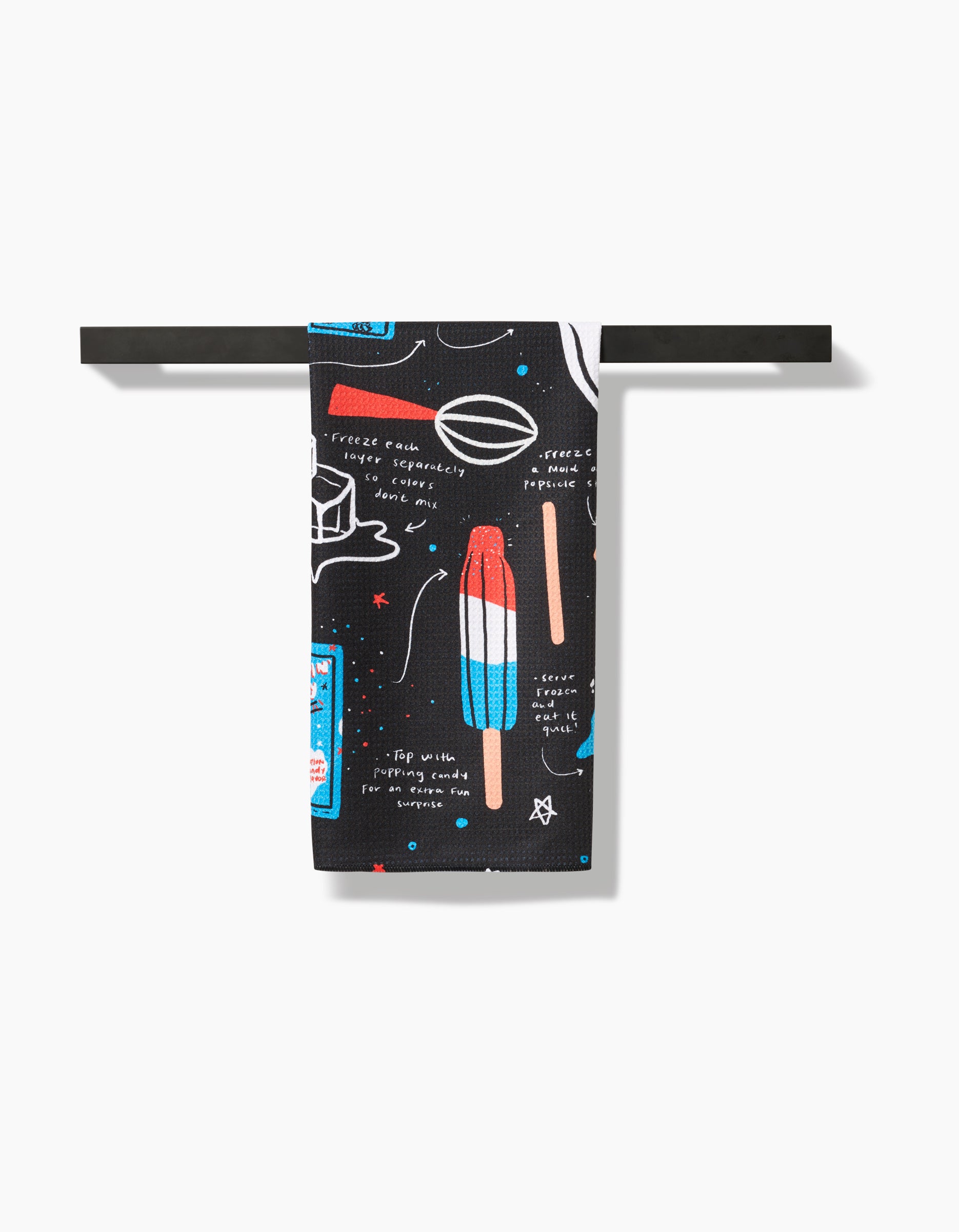 The Popsicles tea towel by Janna Sue Design is black, made from recycled materials, and features colorful illustrations of a popsicle, watermelon slice, and kitchen utensils hanging on a black rod.