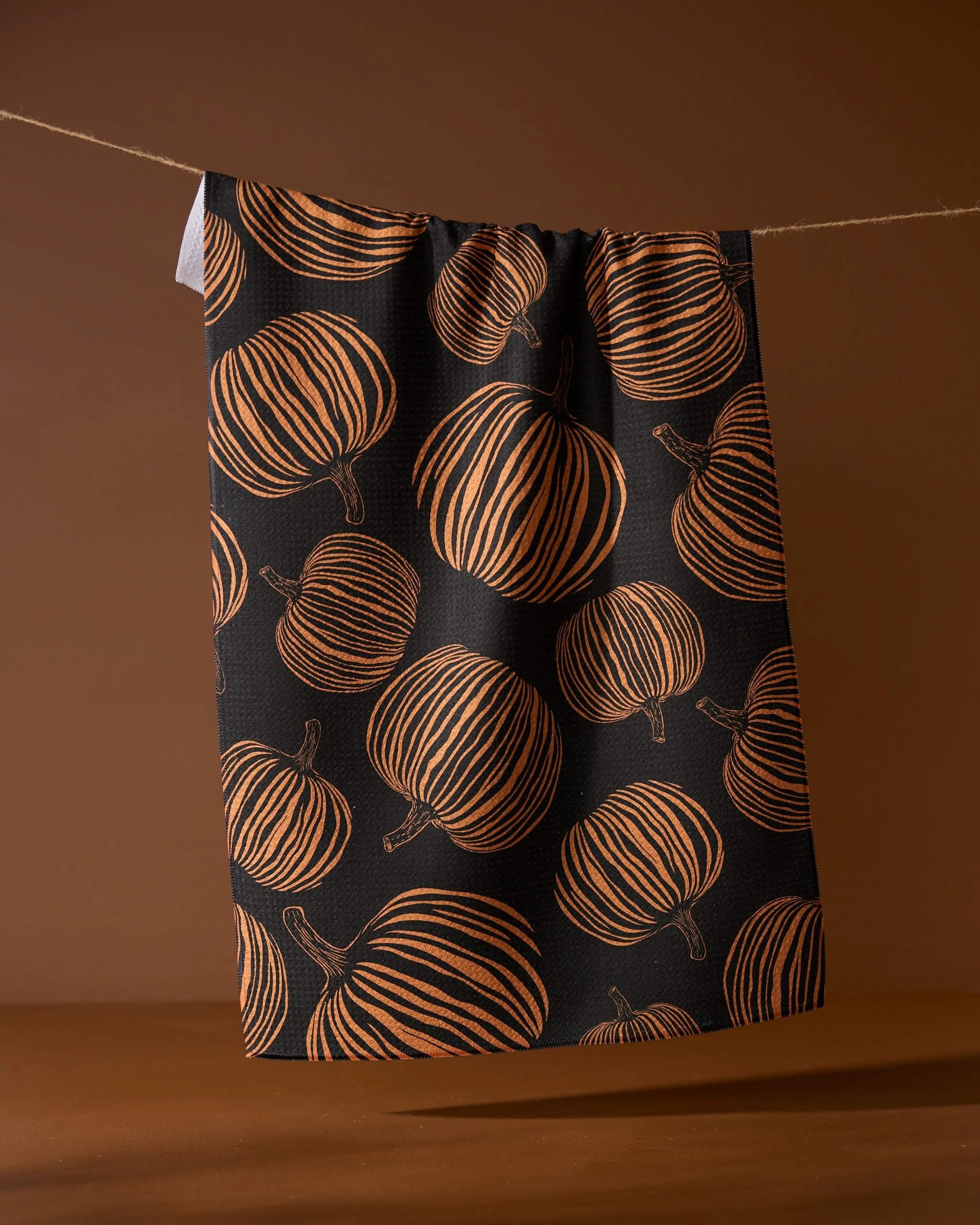 The Geometry Pumpkin Noir recycled kitchen towel, featuring an orange pumpkin pattern on black, hangs from a string against a brown background.