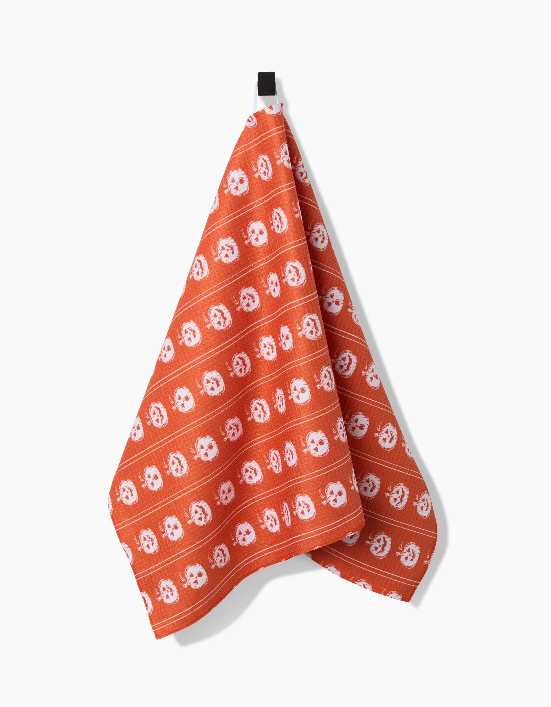 The Pumpkin Pinstripes kitchen towel by Aubrey Fairchild, featuring white smiling faces on orange and made from recycled materials, hangs from a black hook against a plain white background.