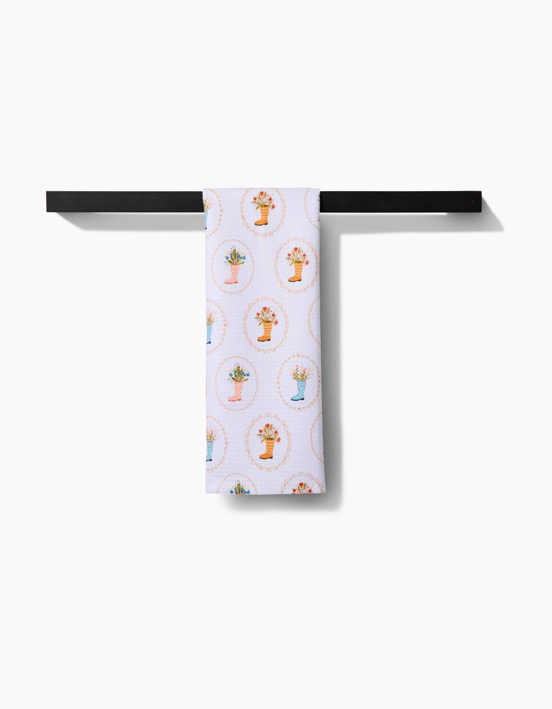 The Sara Mari Rainboots in Bloom tea towel, made from recycled materials, displays colorful potted plant designs and hangs neatly on a horizontal black towel rack against a white background.