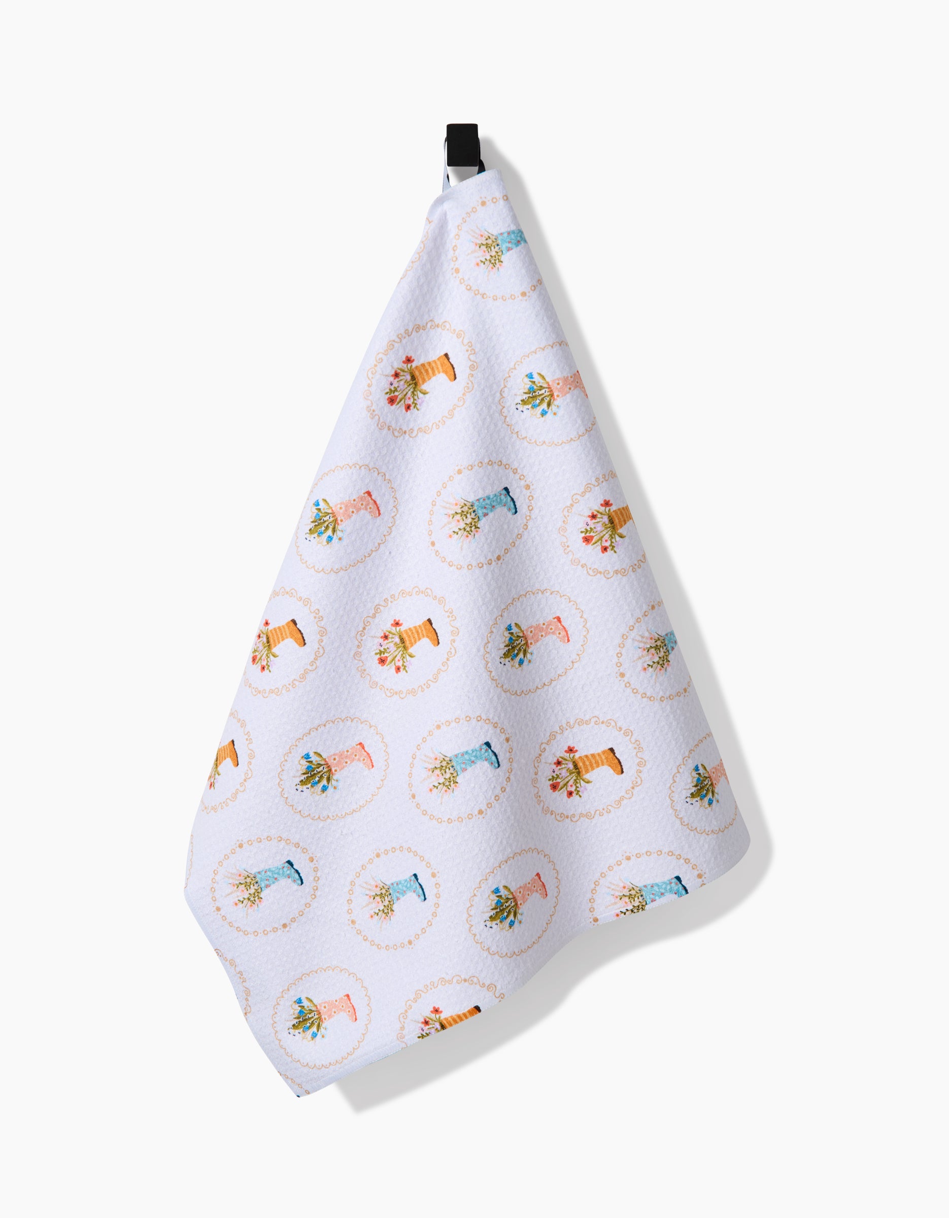 The Sara Mari Rainboots in Bloom tea towel, crafted from Geoweave fabric, features a white background with colorful rainboots filled with flowers, hanging from a black hook against a plain backdrop.