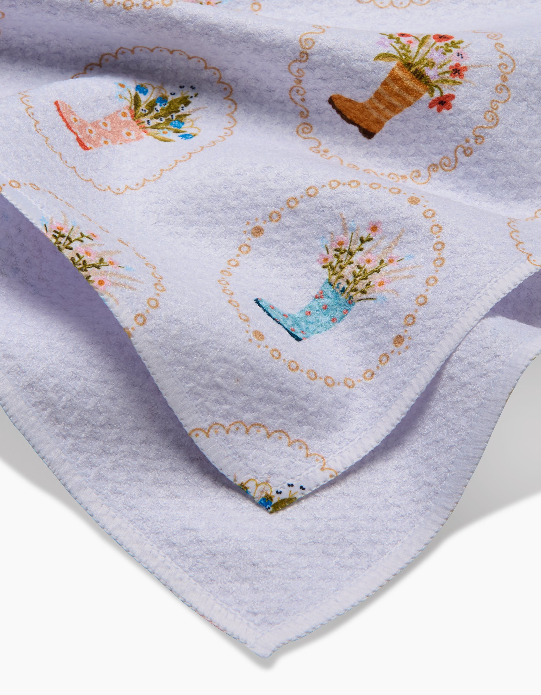 The Rainboots in Bloom tea towel by Sara Mari is a soft, light purple, textured towel made from recycled materials, featuring colorful boots with flowers and gold swirls in a charming repeating pattern.